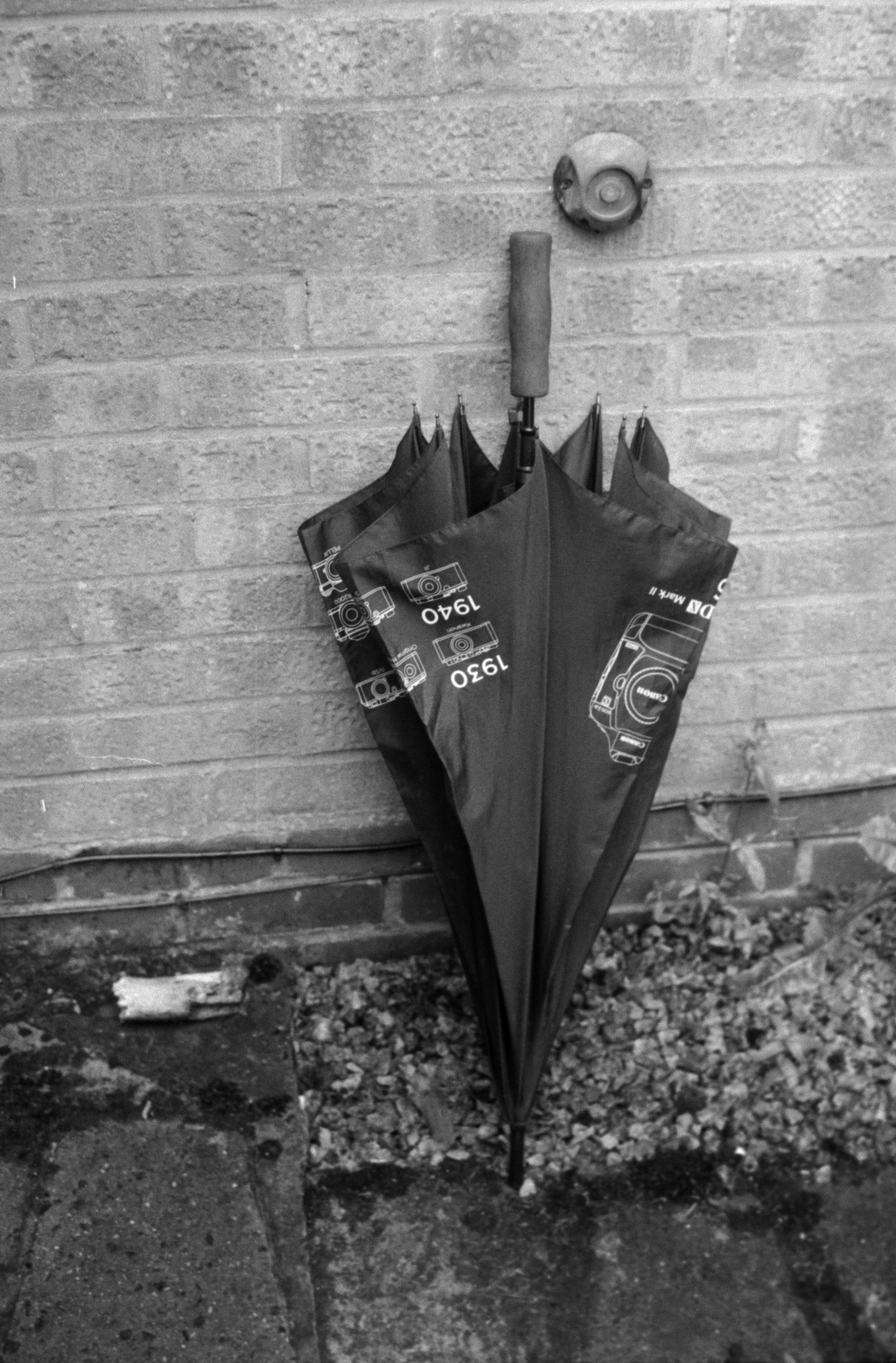 A closed umbrella with a pattern of automotive diagrams, standing upright against a brick wall.