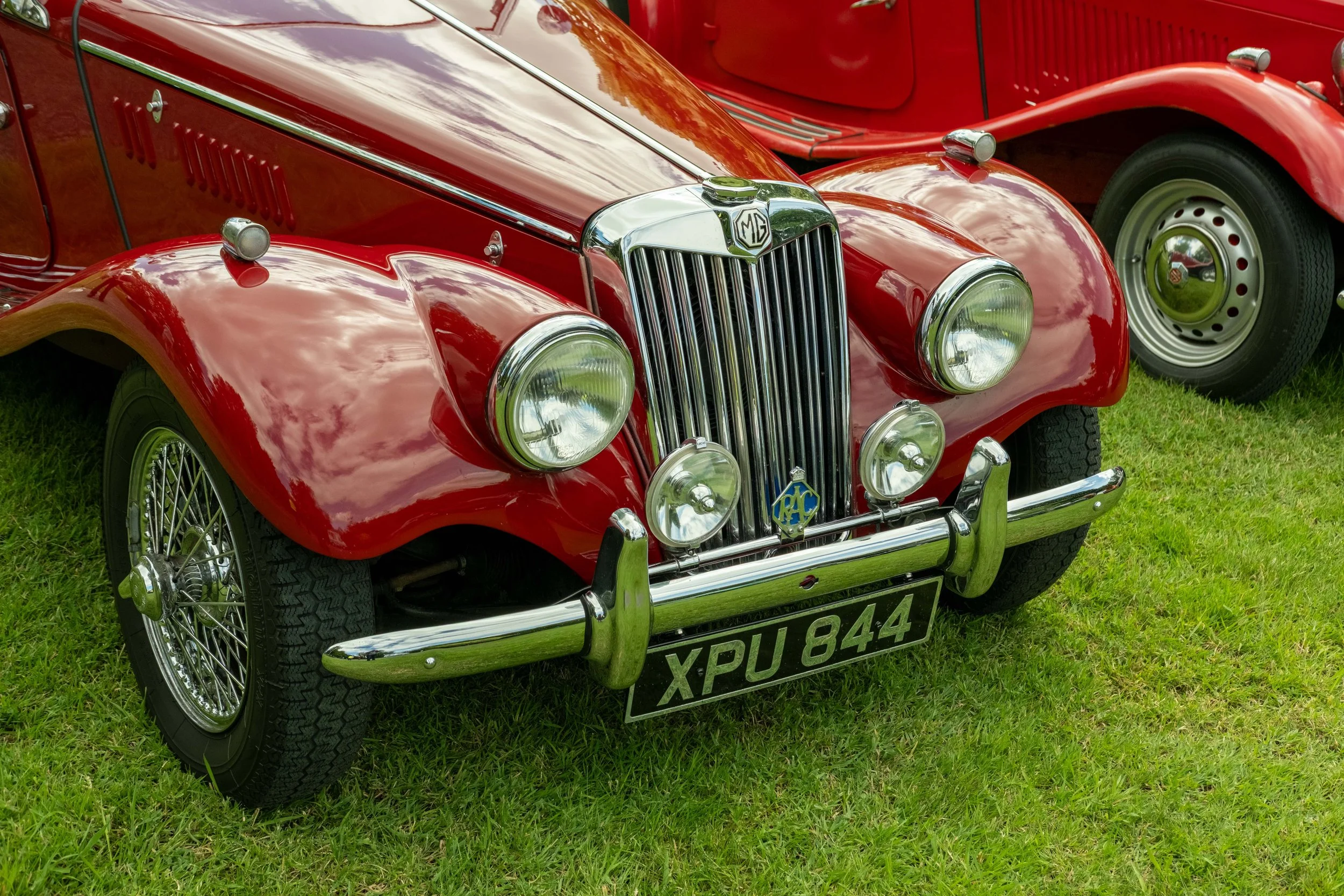 A close-up of a vintage red MG convertible car parked on grass, showing front grille with MG emblem, round headlights, chrome bumpers, and wire-spoke wheels.