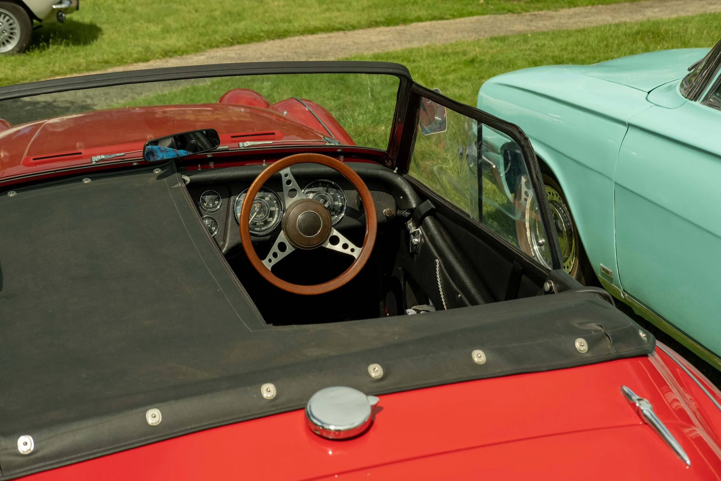 Close-up of the interior dashboard and steering wheel of a vintage red convertible car, with part of a teal-colored car visible next to it on a grassy area.