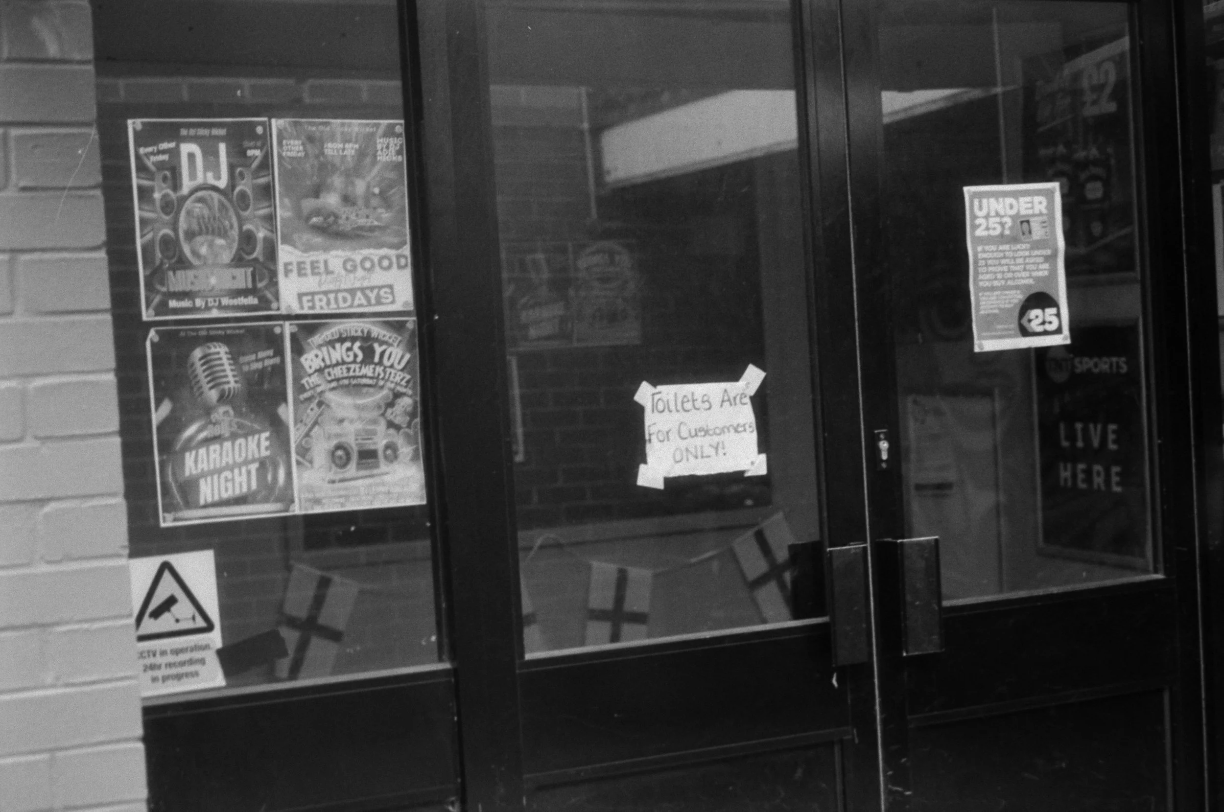 Black and white photo of store window with posters, including a sign that reads 'Toilets Are For Customers ONLY!' and another about being under 25, with a brick wall visible on the side.