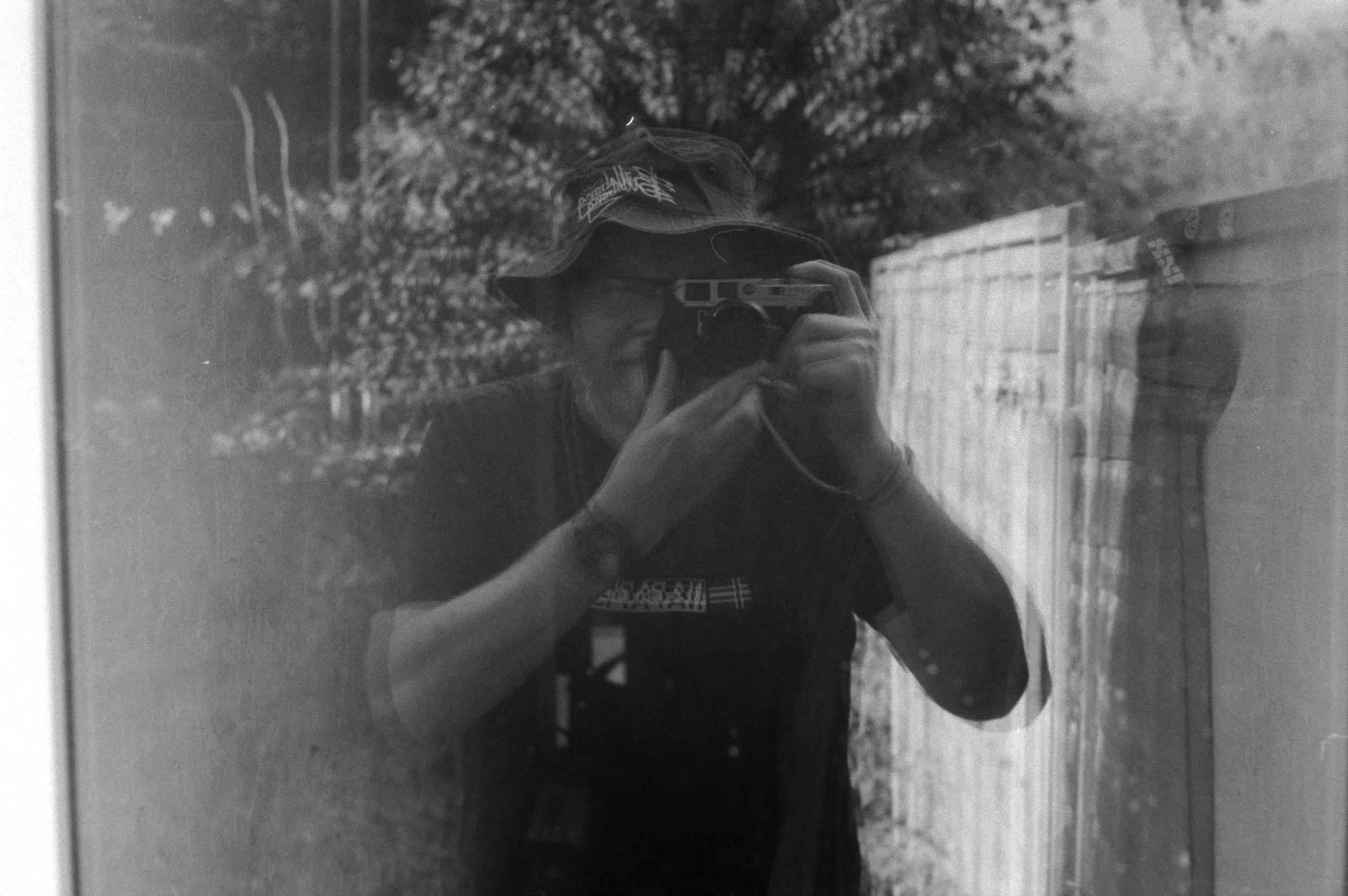 Black and white photo of a person taking a selfie in a window or mirror, with trees and sky reflected in the background.