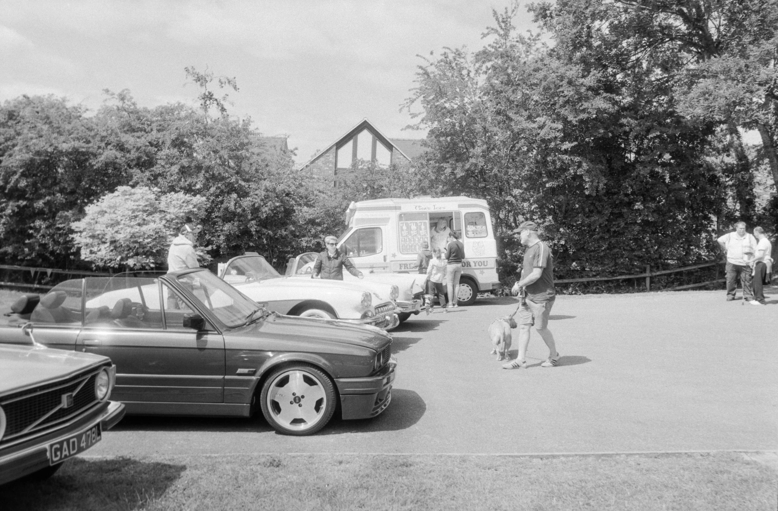 People standing and walking near parked cars and a food truck in an outdoor setting, with trees and a building with triangular windows in the background.