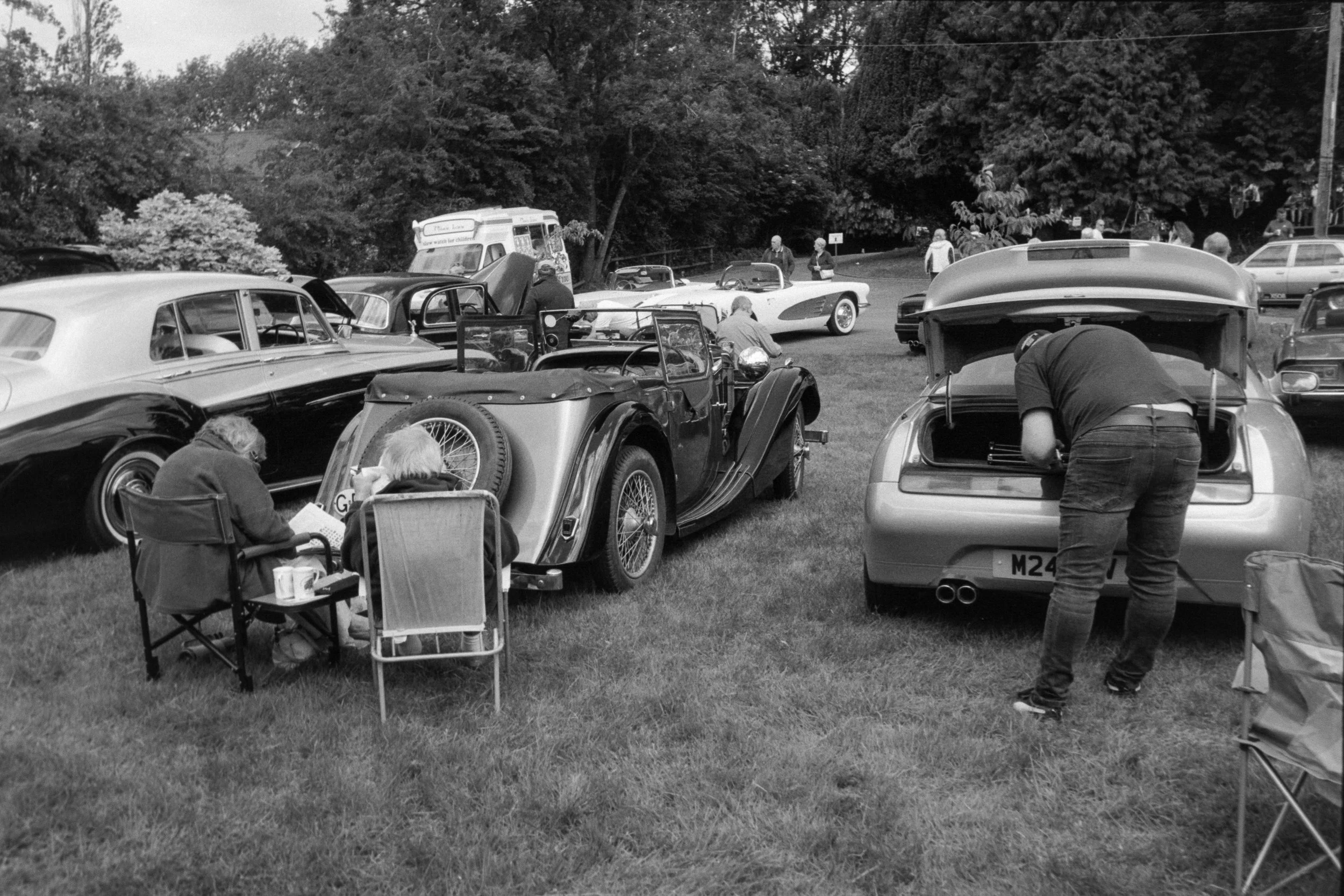 A black-and-white photo of a vintage car show in a park, with classic cars, people examining cars, and trees in the background.