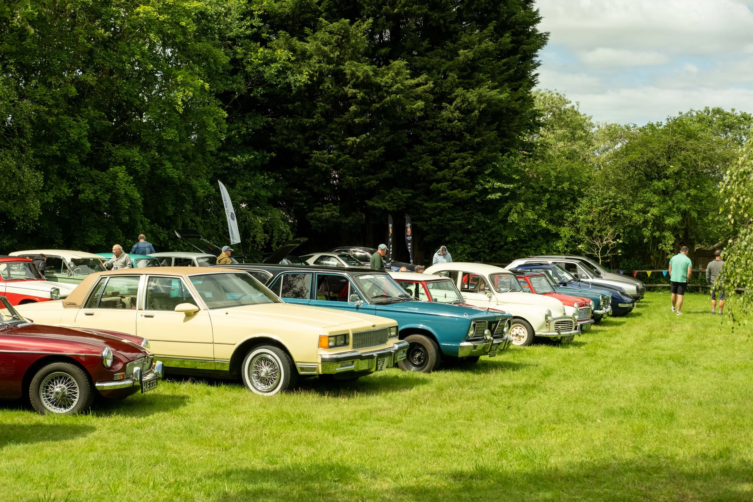 A vintage car show with various classic cars parked on a grassy field surrounded by tall trees under a cloudy sky, and people walking around inspecting the cars.