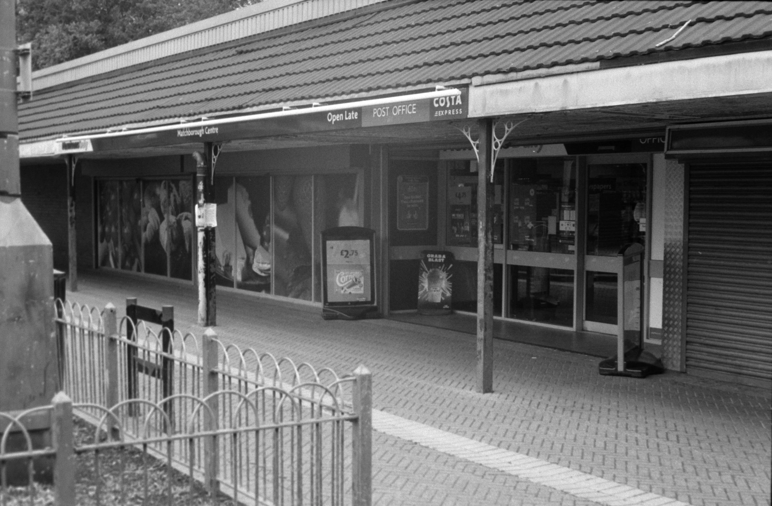 Black and white photo of a small shop with shopfront signage, including a Coca-Cola vending machine outside. The shop has large display windows, and there is a covered porch with metal support posts. A low fence lines the sidewalk in front.