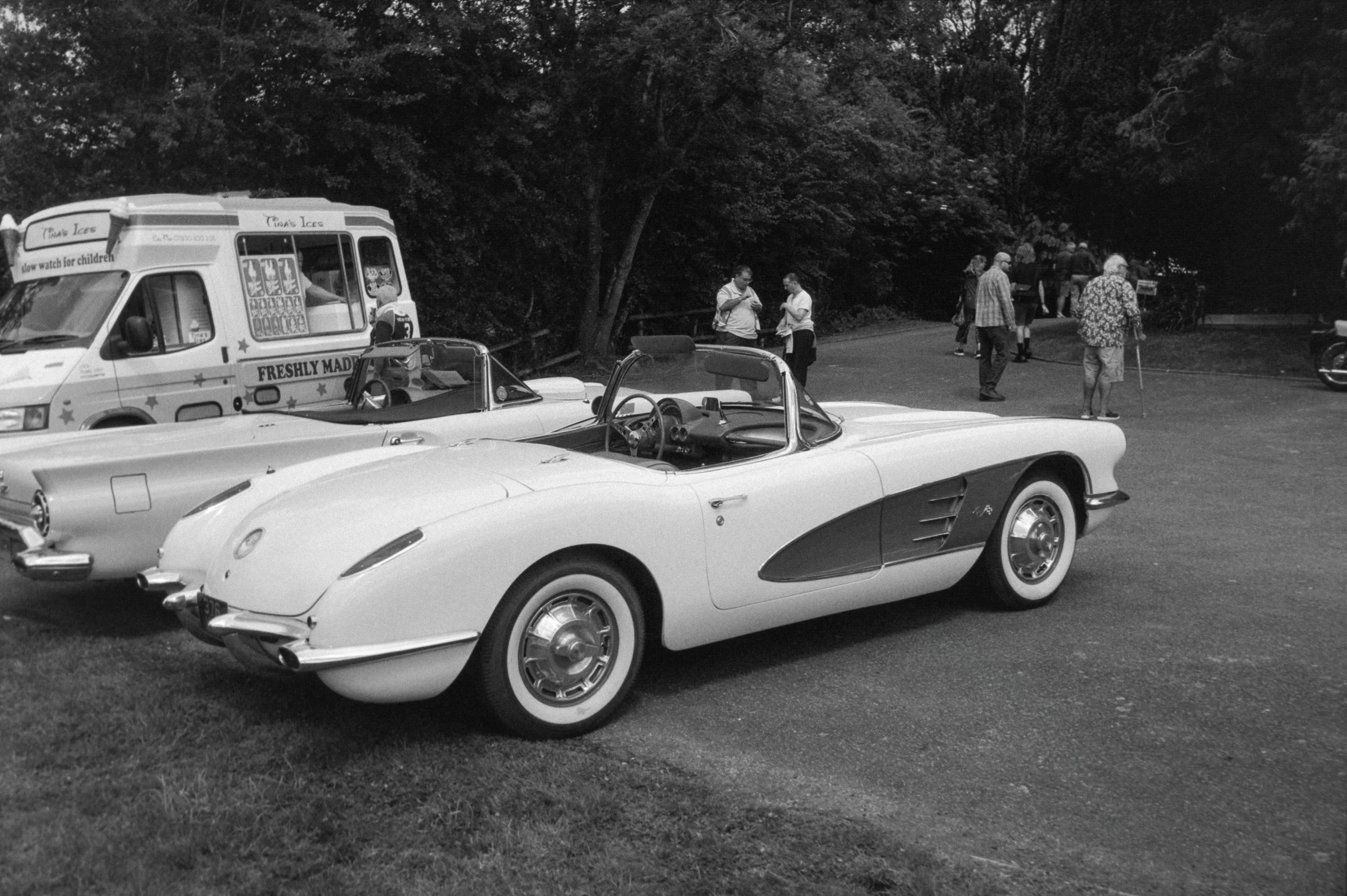A black and white photo of a vintage scene with a classic white convertible car in the foreground, a vintage ice cream truck on the left, and several people walking and standing near a wooded area in the background.