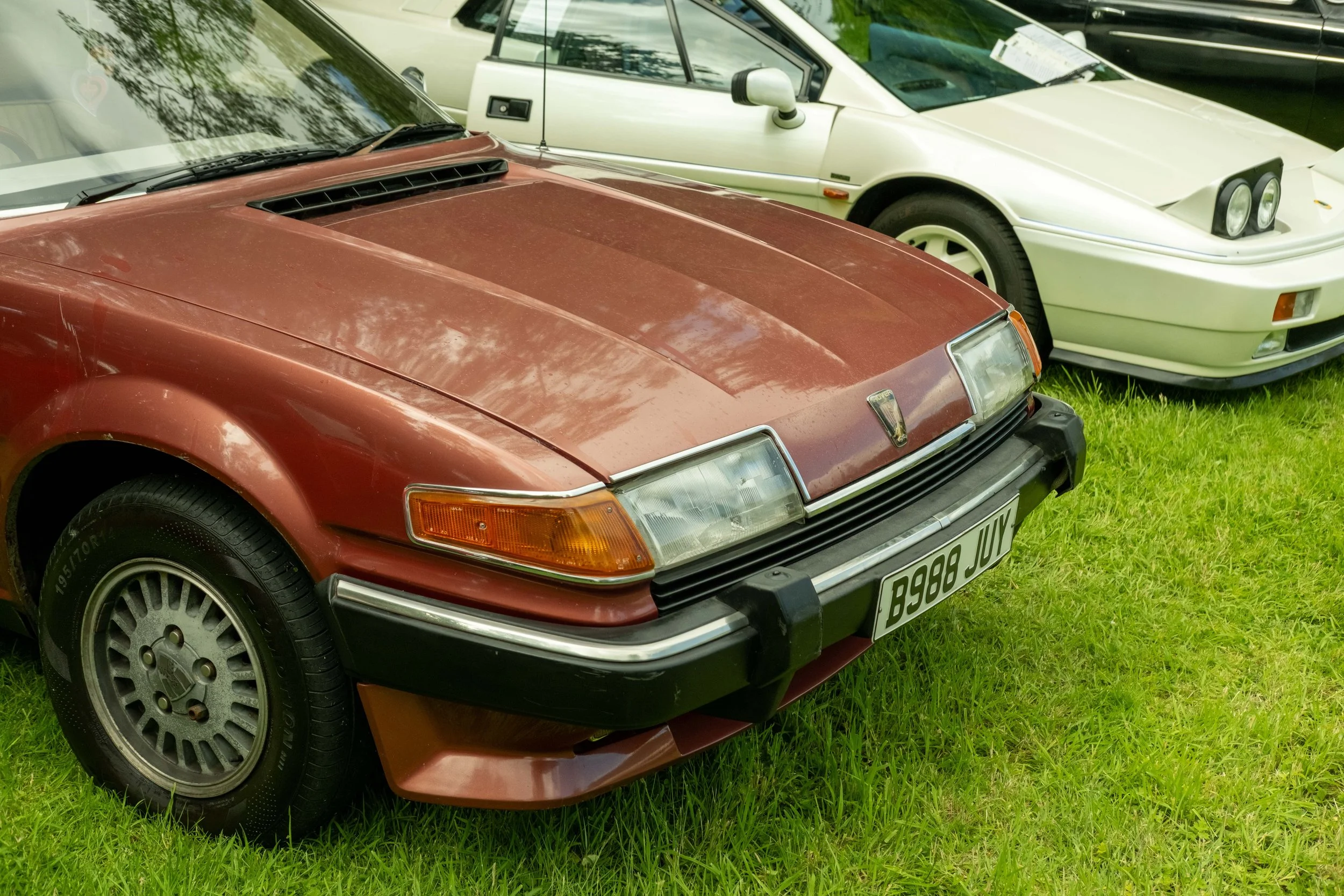 A close-up of a red vintage sports car with black tires parked on green grass, showing the front and part of the side. The car appears to be a classic model from the 1980s.
