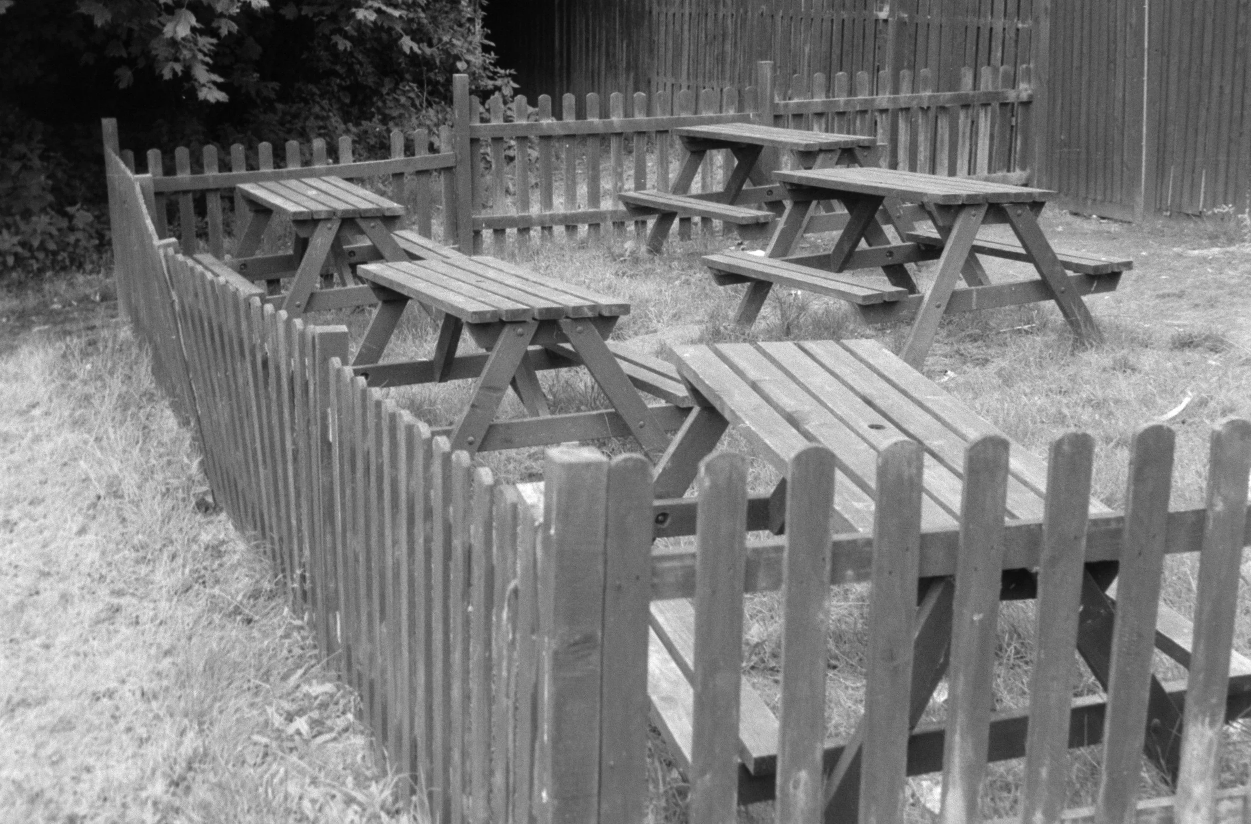 A small fenced area containing a wooden picnic table with attached benches, situated on a grassy lawn with bushes and a wooden fence in the background.