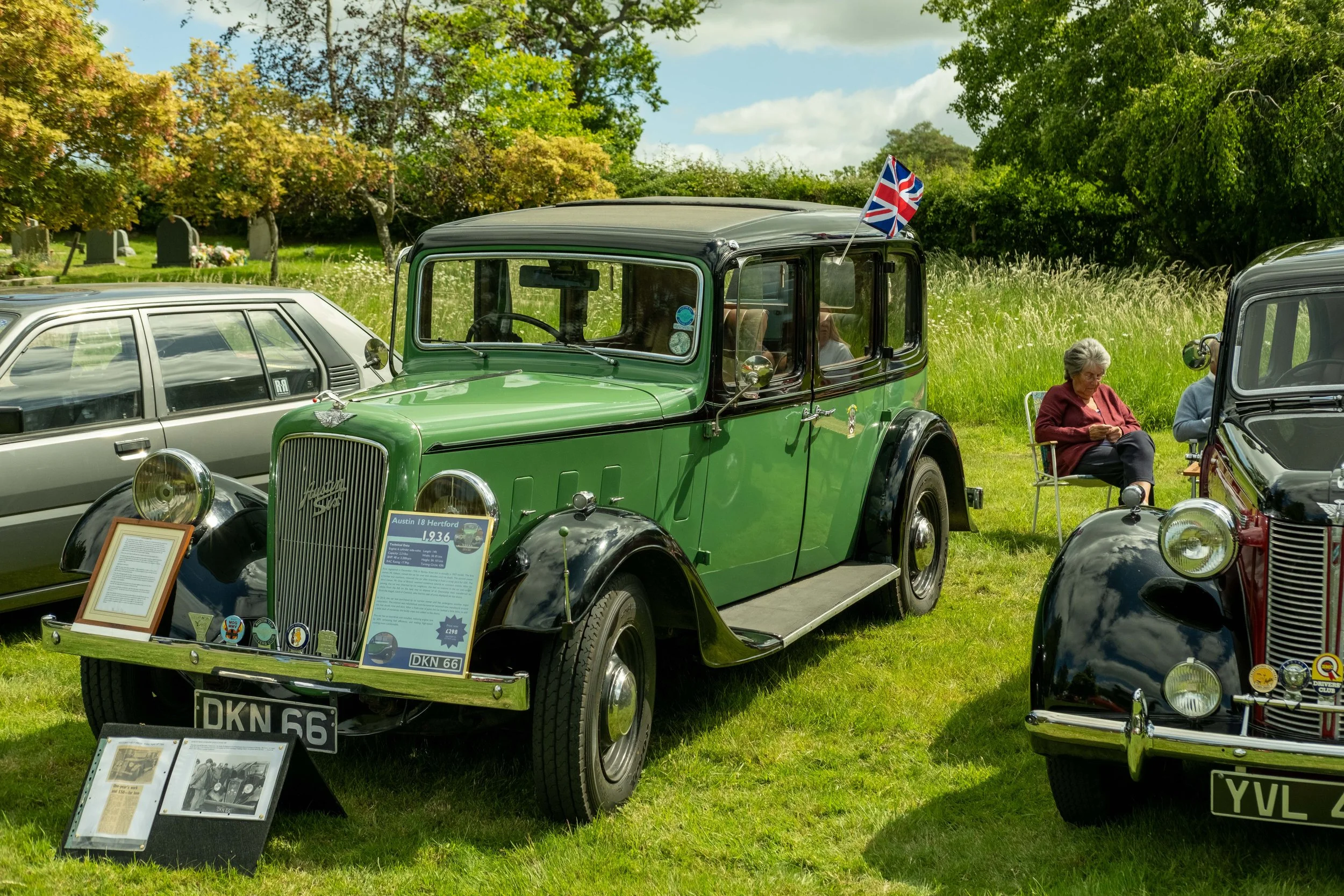 A green vintage car displayed at a classic car show, with a Union Jack flag on the roof, surrounded by other antique cars, trees, and a grassy field.