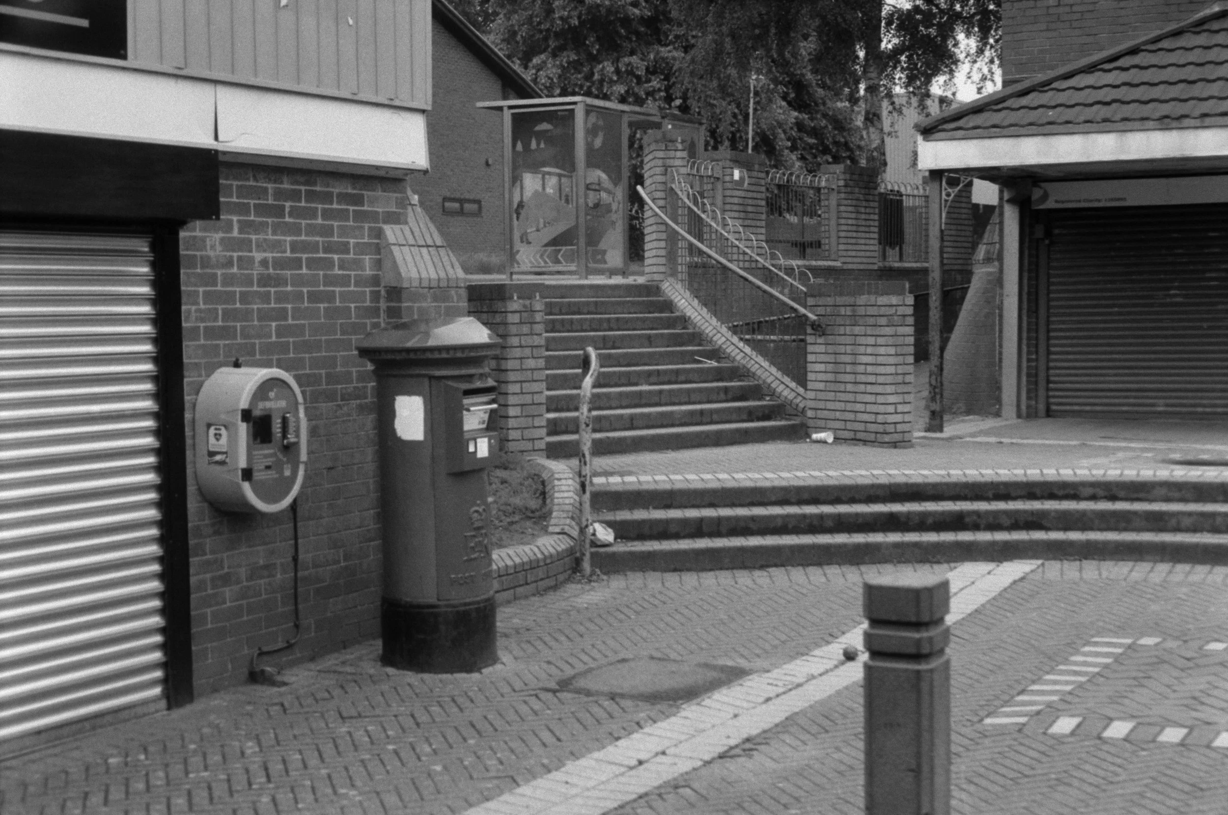 A black and white photo of an outdoor staircase with brick steps and metal railings, leading up to a gate in a residential area. There are two trash bins and an electrical box near the bottom of the steps and a building with brick and siding walls ne