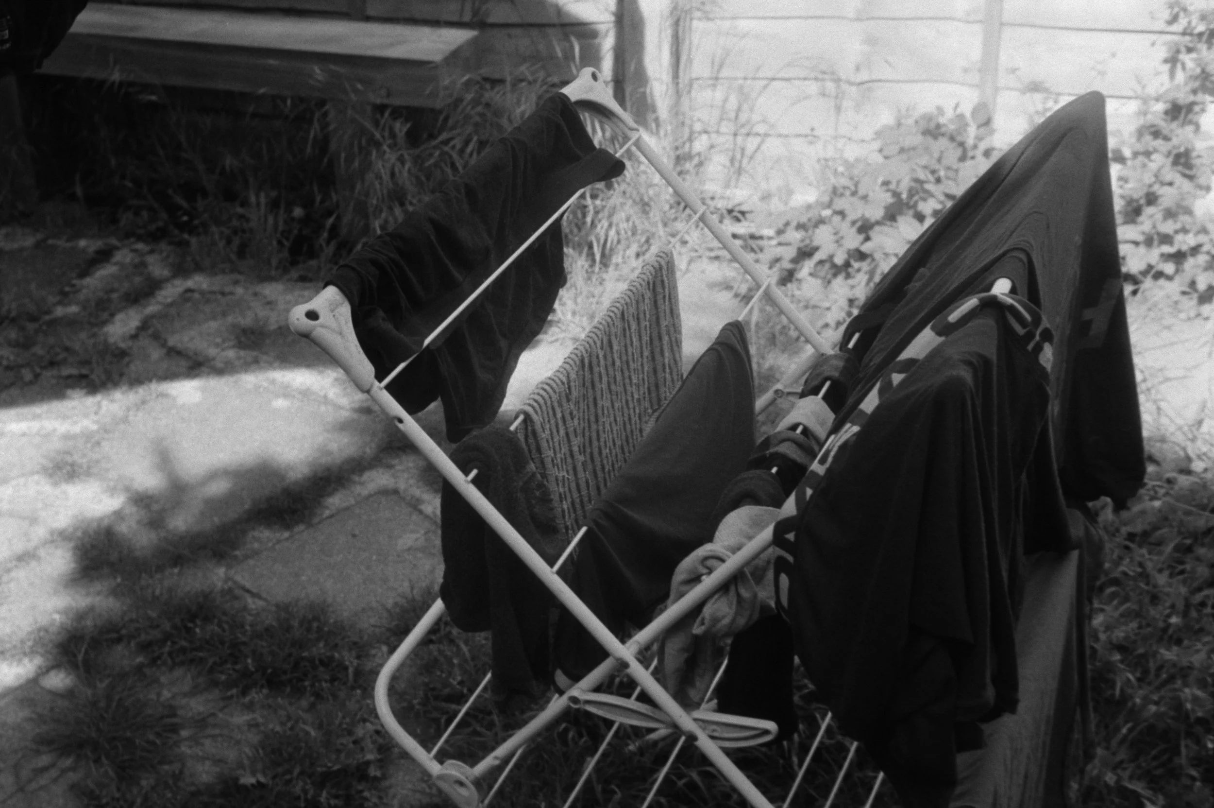 A black and white photo of a person lying on the ground in a lawn chair outdoors. The person's legs are visible, and they are wearing dark clothing. The lawn chair is placed on the grass beside a pathway. In the background, there are plants, a wooden
