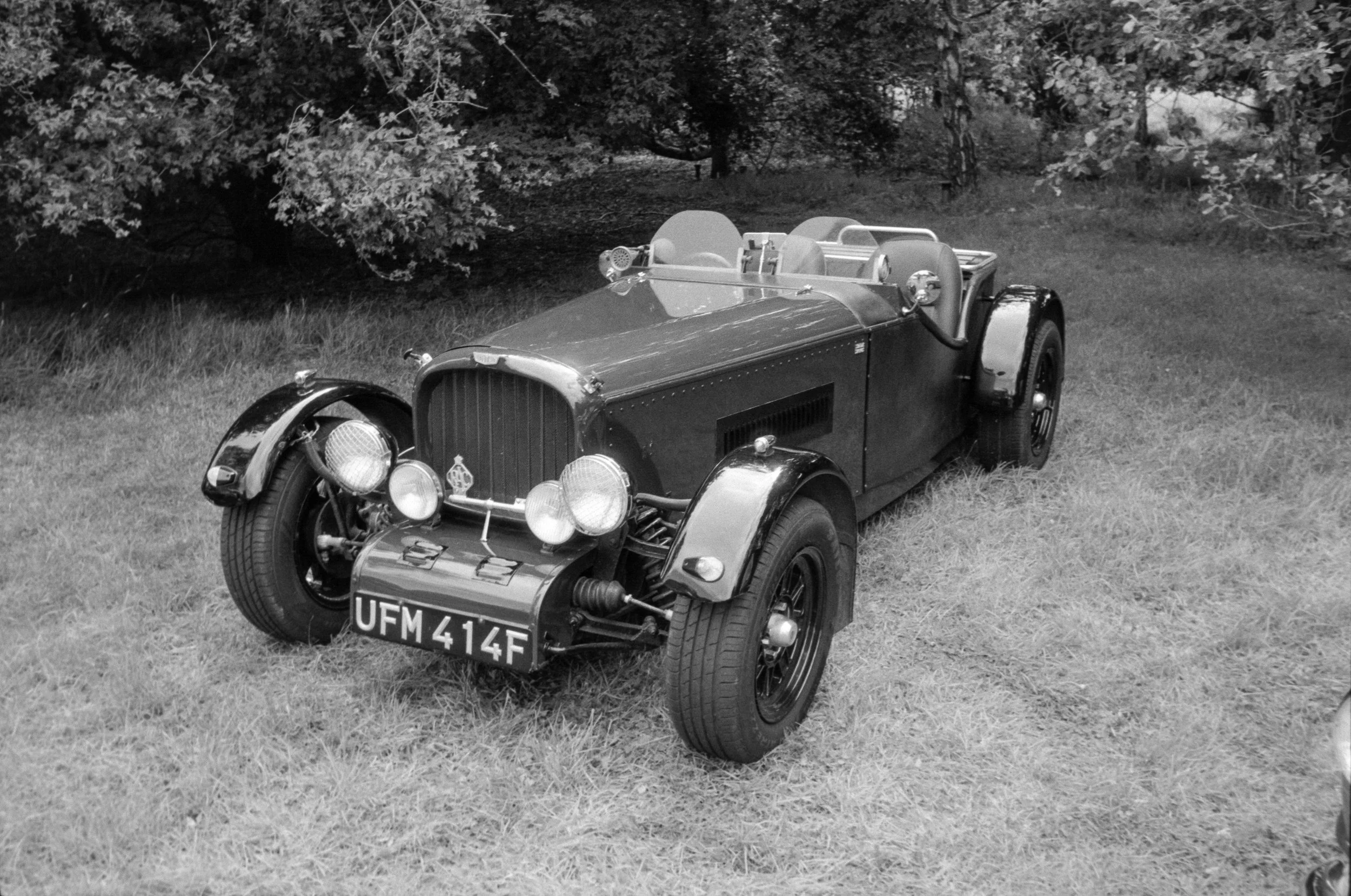 A vintage open-top sports car parked on grass, with trees in the background in black and white.