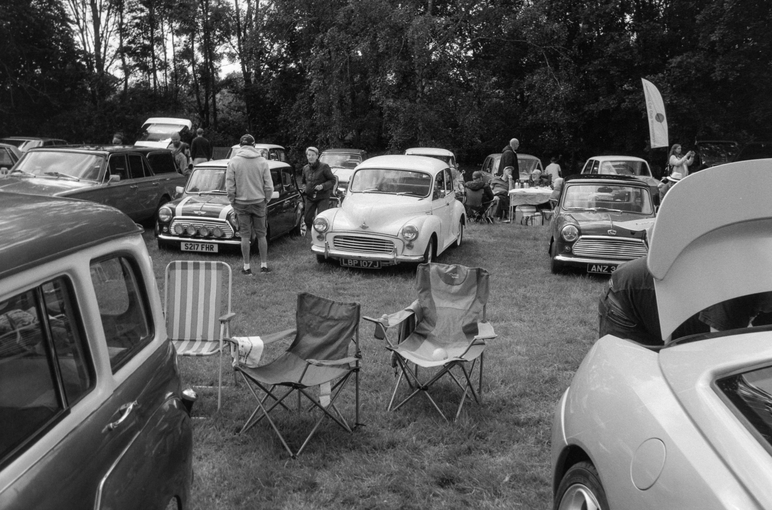 A gathering of people outdoors around vintage cars and seated in camping chairs in a park-like setting.