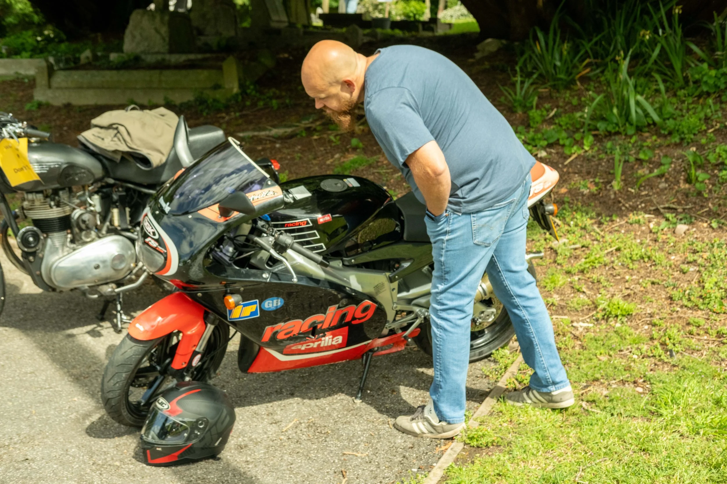 A man with a bald head and beard is bending over a black and red Aprilia racing motorcycle, inspecting or working on it. He is wearing a blue t-shirt, jeans, and sneakers. Nearby on the ground is a black helmet with a red design, and a beige helmet i
