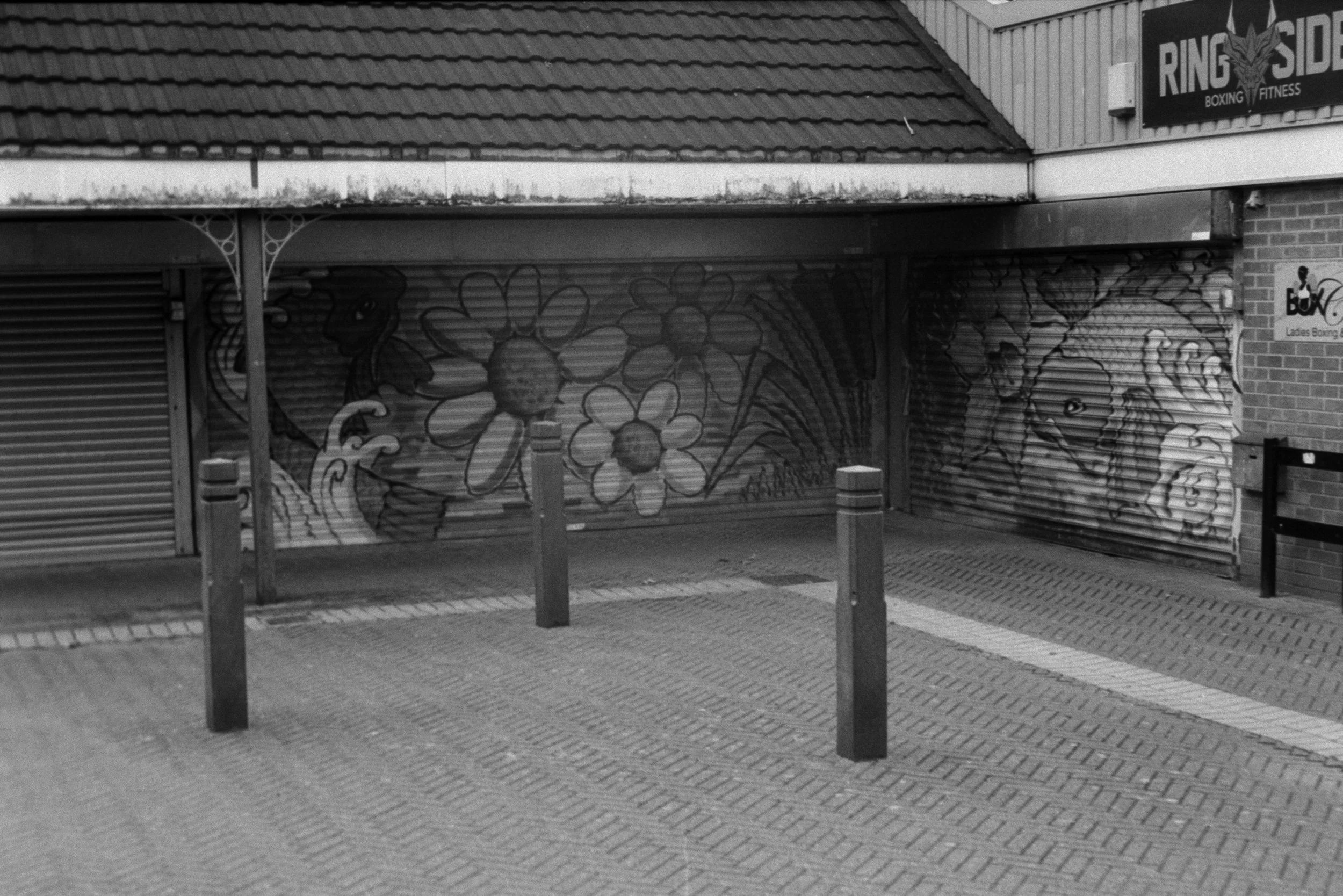 Black and white photo of closed graffiti-covered metal shutters in front of a brick building. There is a sign that reads 'Ring Side Boxing Fitness' and a small poster for Ladies Boxing on the brick wall to the right.