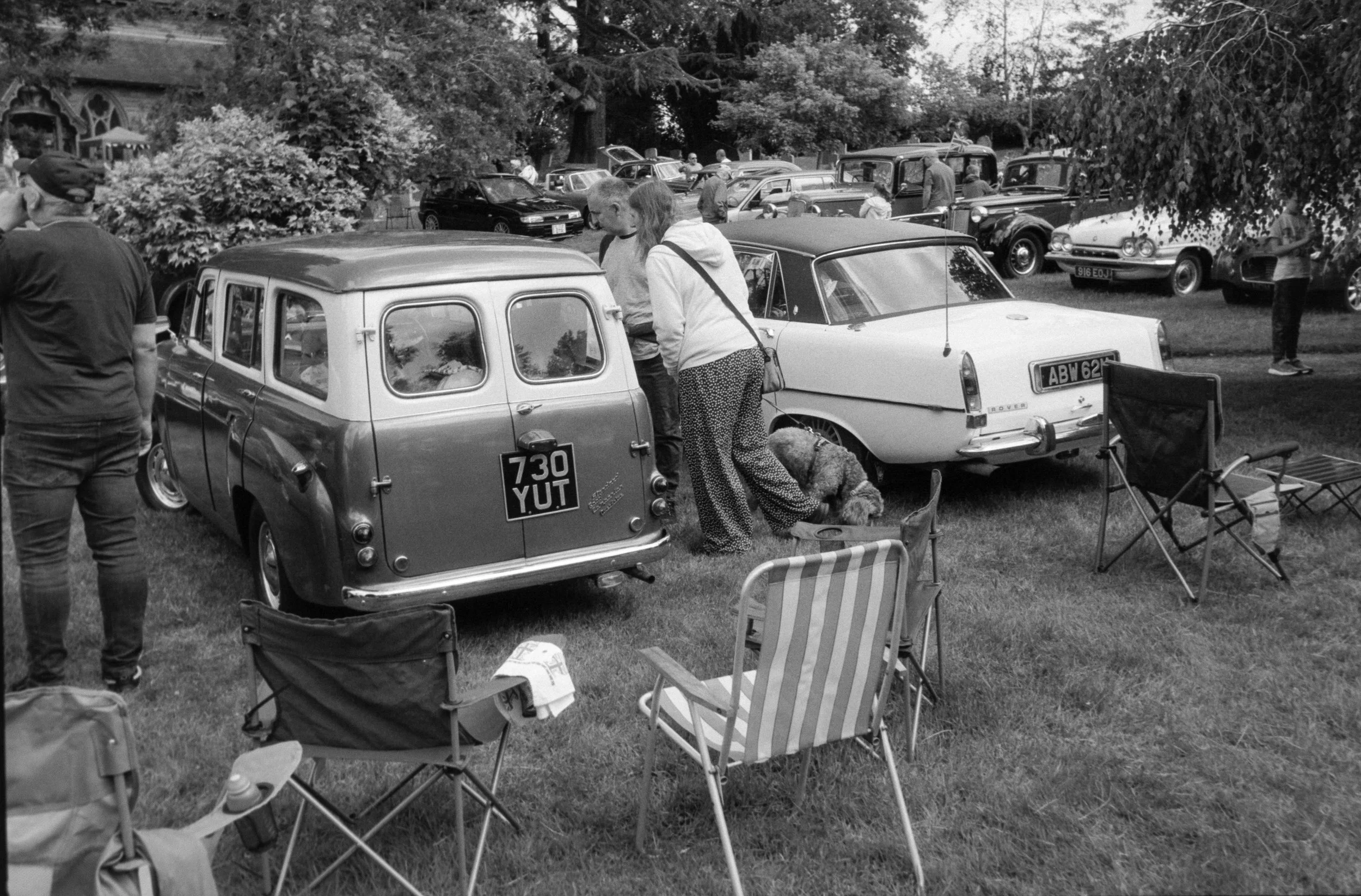 Black and white photo of a car show in a park with people walking around and looking at vintage cars. Folding chairs are set up on the grass.
