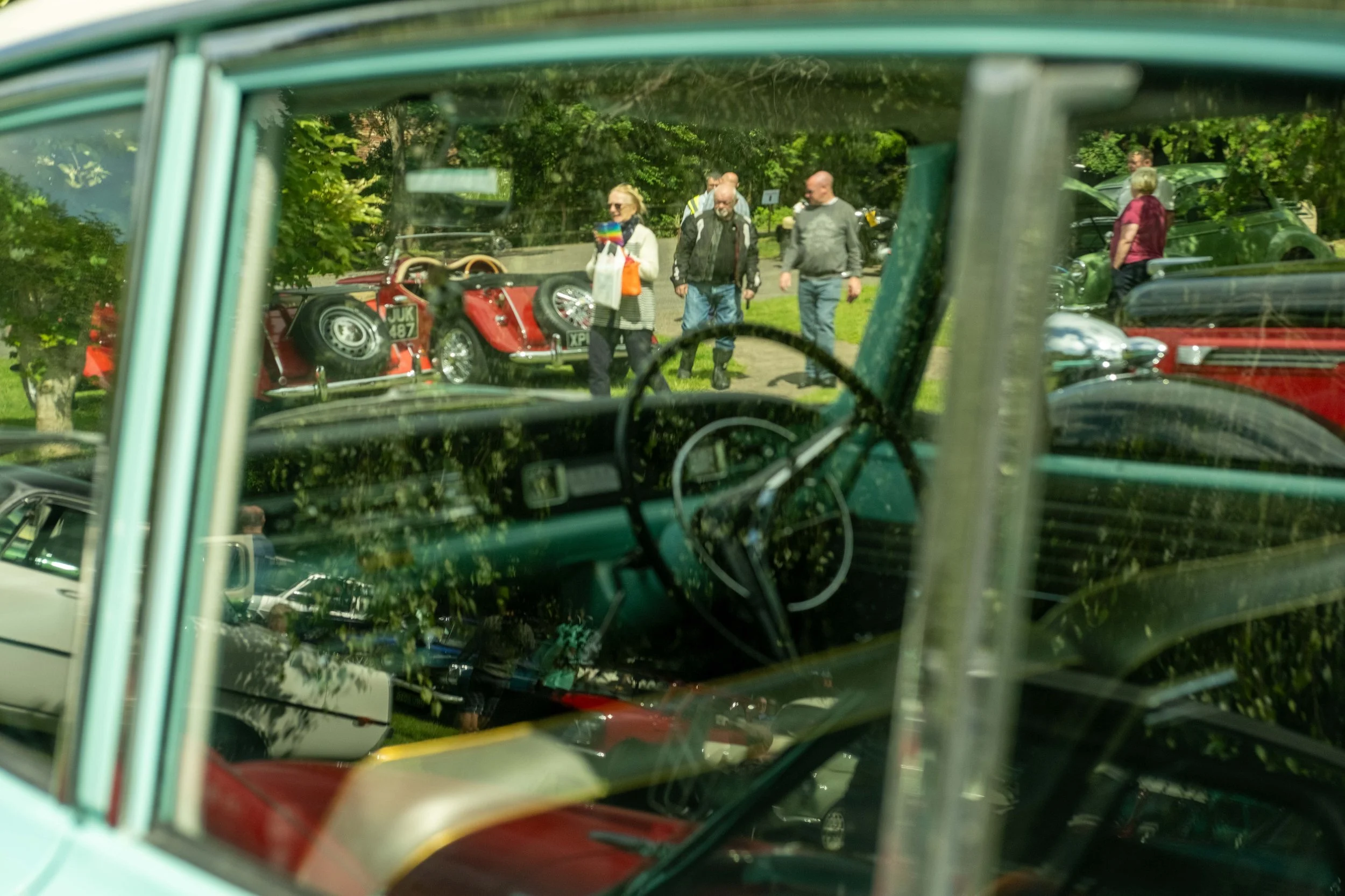 View through a car window showing people walking and vintage cars on a grassy area in a park, trees in the background.
