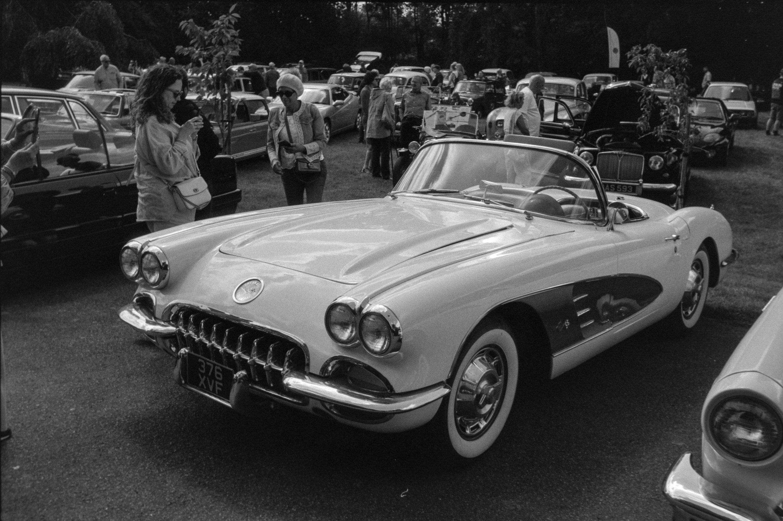 A black and white photo of a vintage car show. The foreground features a classic convertible with distinctive front grille and dual headlights. People are gathered around, some taking photos, others talking. In the background, many other classic cars