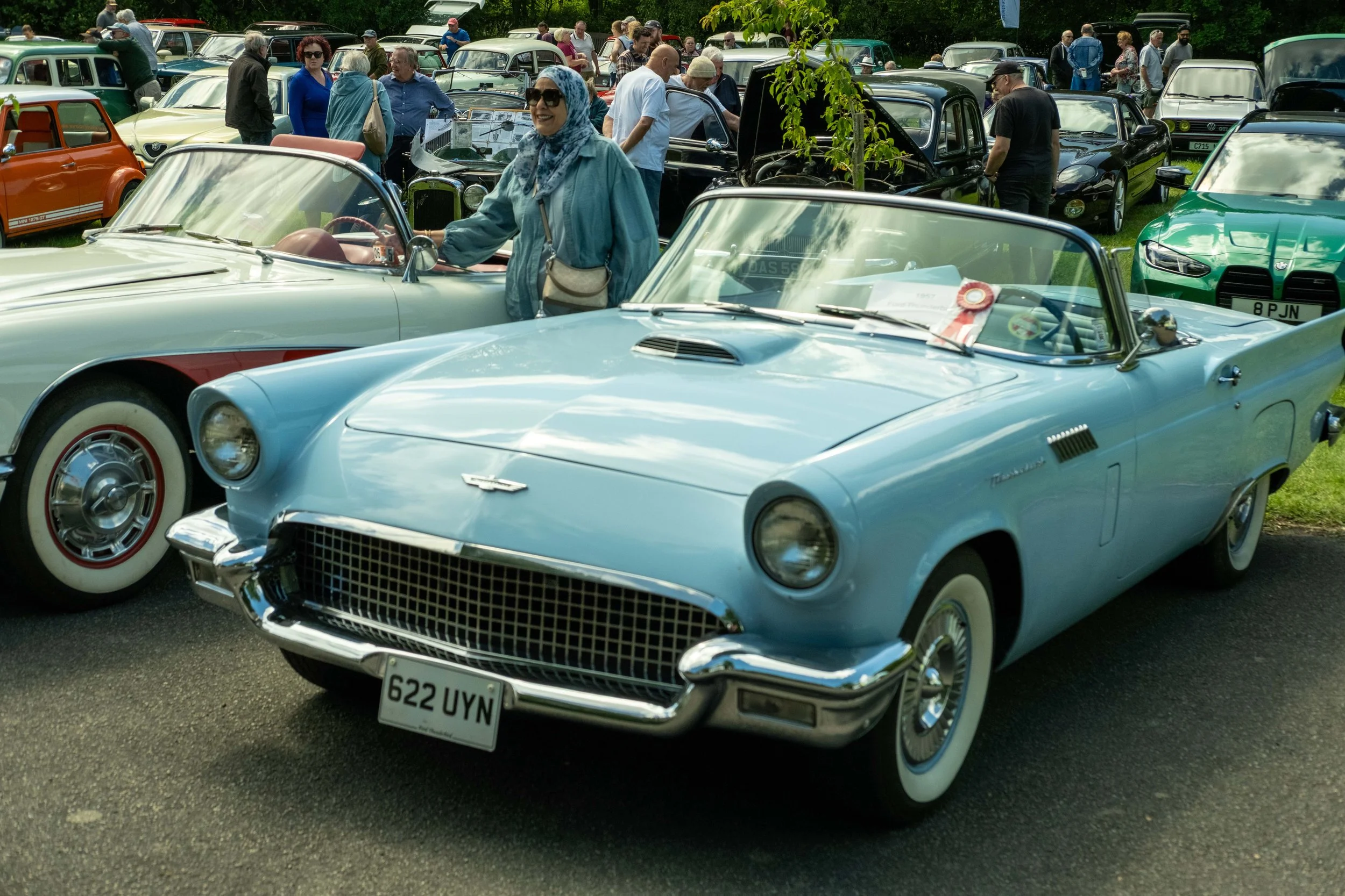 A woman wearing sunglasses and a headscarf stands beside a vintage blue convertible car at a car show, surrounded by other classic cars and people examining vehicles outdoors.