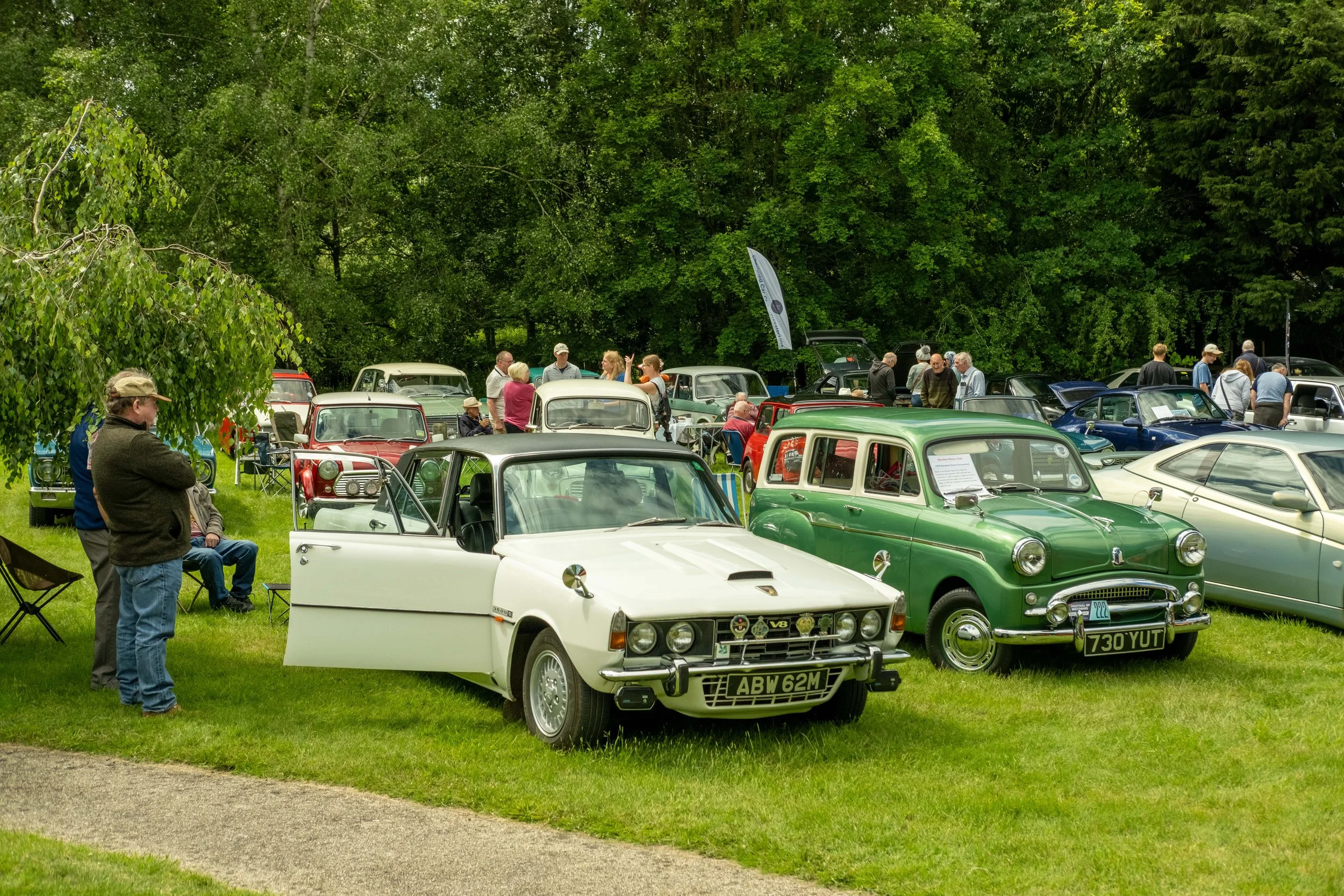 Vintage cars on display at an outdoor car show with people walking and viewing the vehicles among trees.