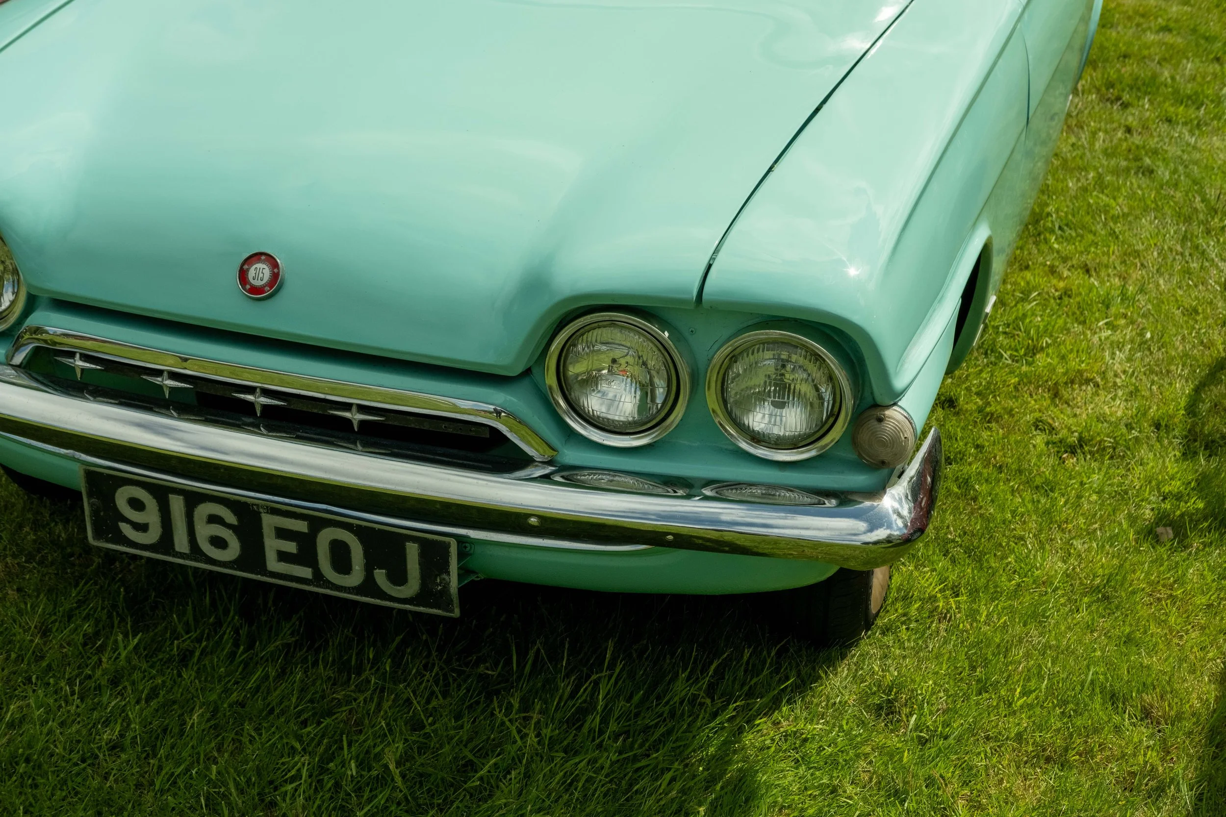 A vintage light blue car, possibly a classic model, parked on grass. The front grille, two round headlights, a small emblem with the number 315, and a black license plate with white characters are visible.