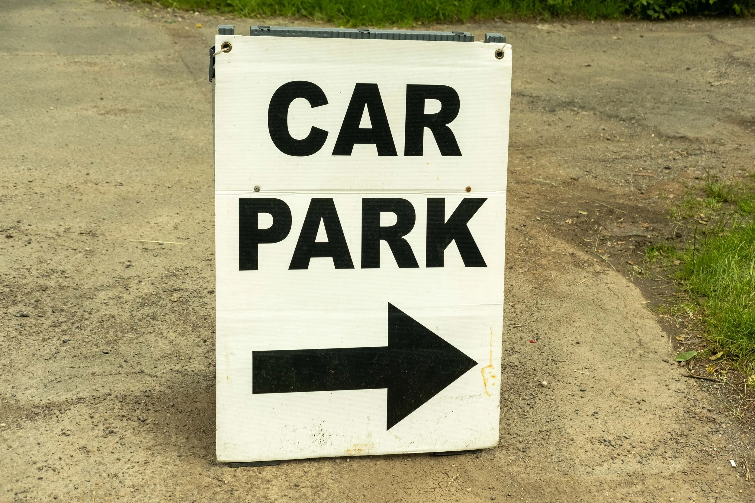 A white sign with black letters reading "CAR PARK" and a black arrow pointing to the right, placed on a dirt ground.