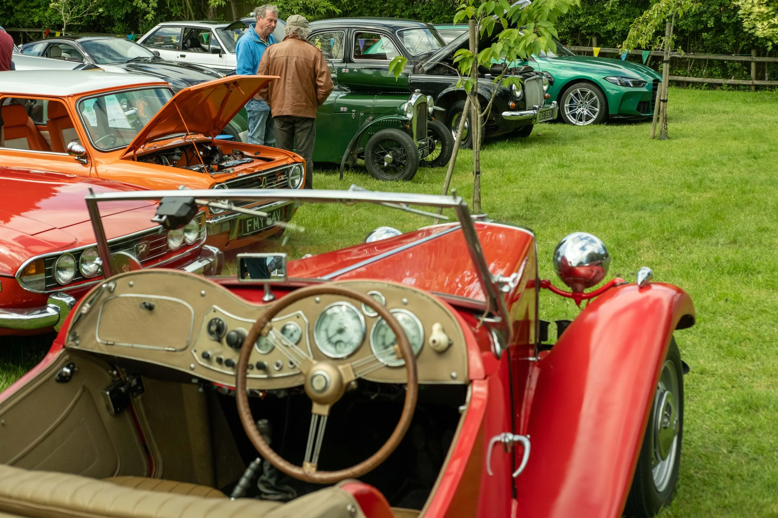 A vintage red convertible car with an open hood displaying its engine, seen from inside with a beige interior and steering wheel, at a classic car show with several other vintage and modern cars parked on green grass, and people walking and talking i