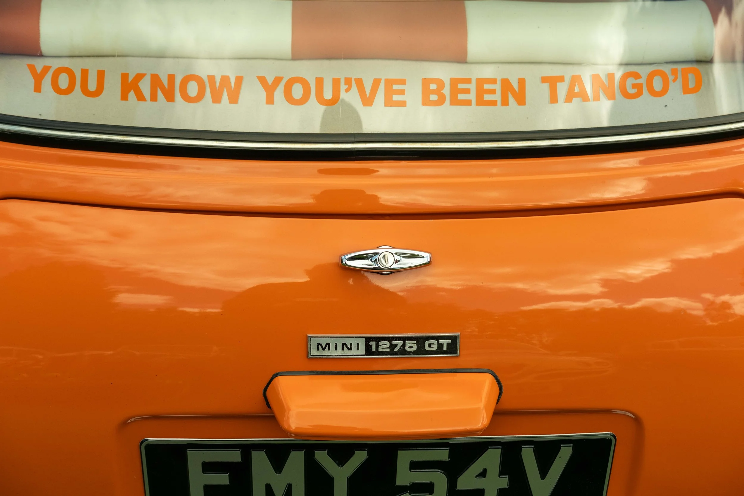 Close-up image of the front of an orange Mini Cooper car, showing the Mini emblem, a license plate reading 'FMY 584V,' a badge indicating it is a 1275 GT model, and a sticker with the phrase 'You know you've been tango'd' on the windshield.