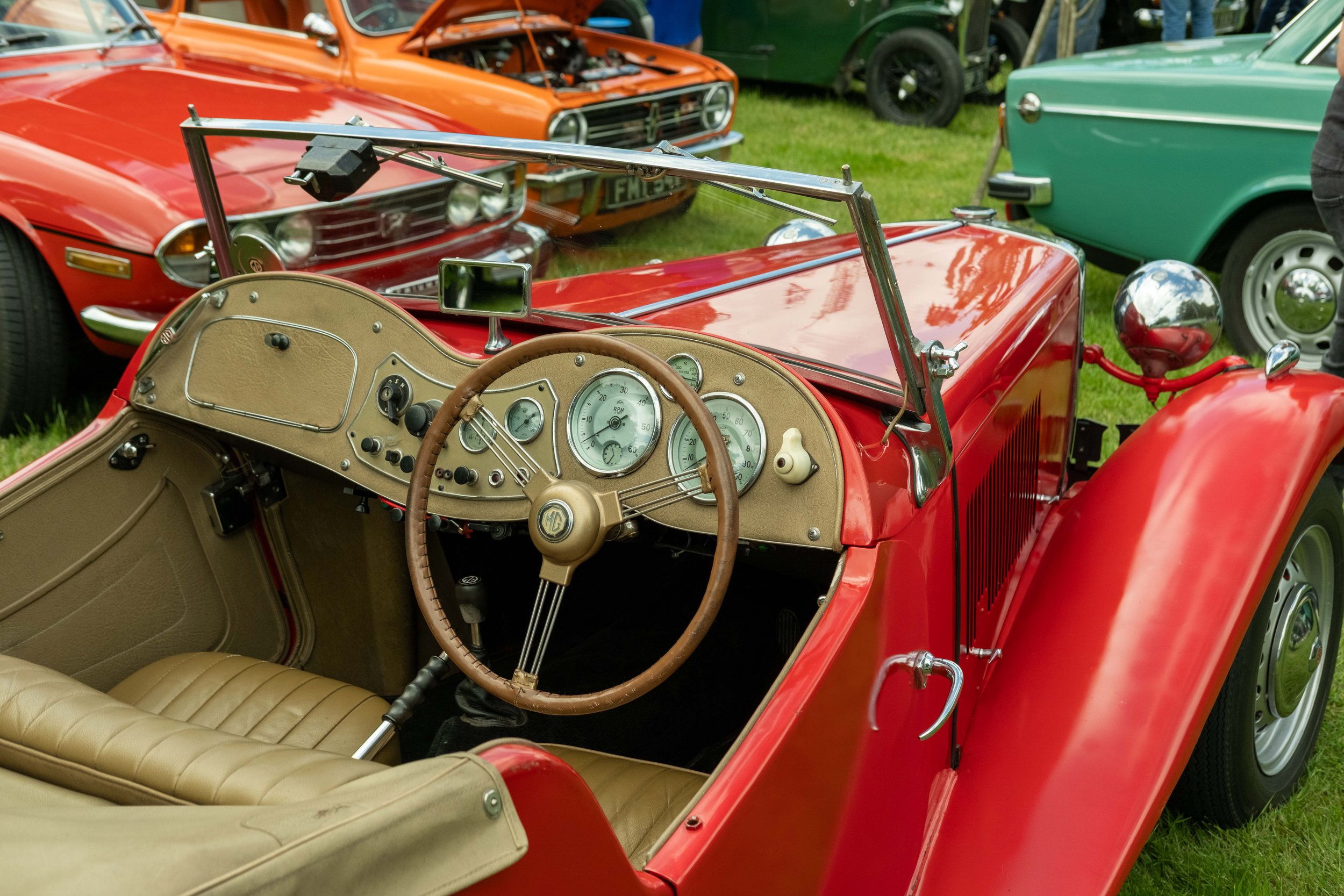 Red vintage MG convertible car with tan interior, parked among other classic cars at a car show.