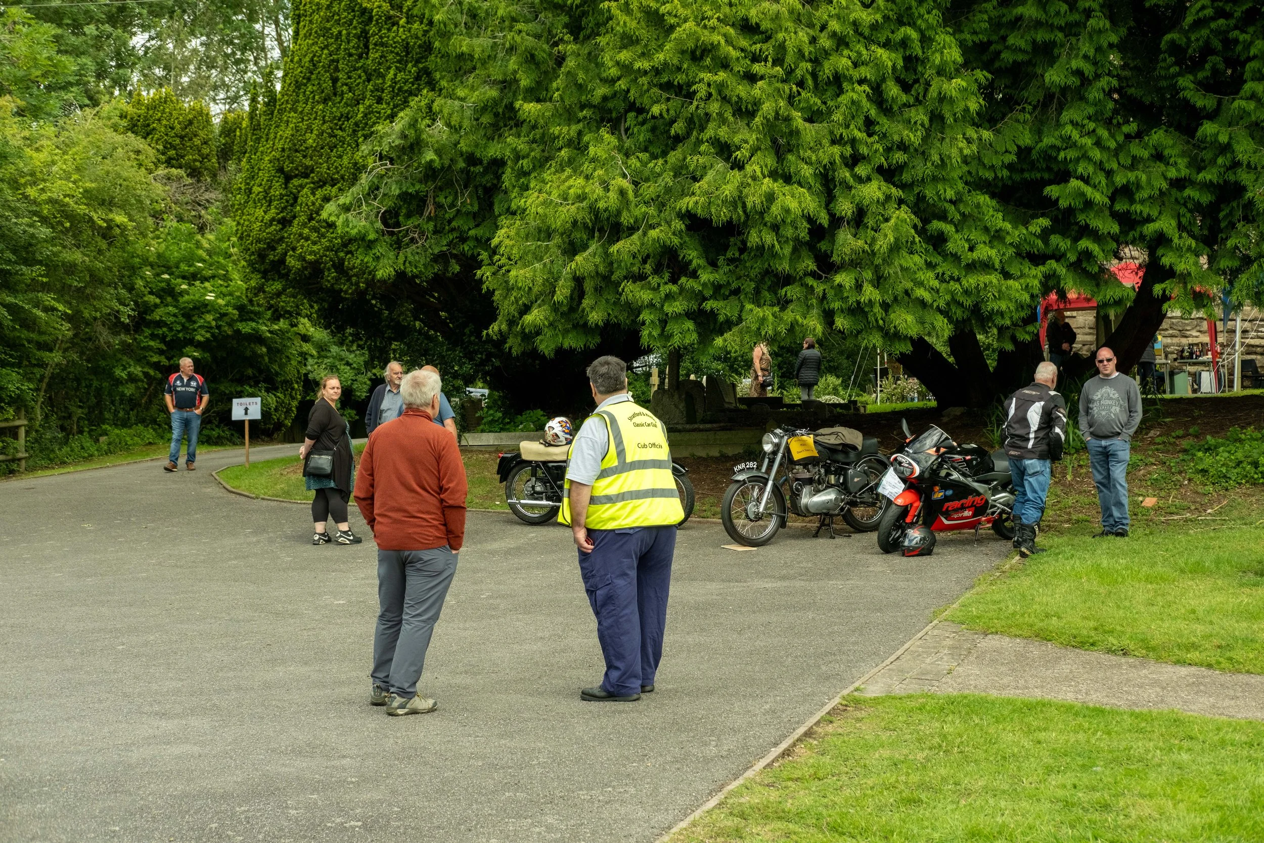 People gather in a park under a large green tree, with some motorcycles parked nearby and a small man-made sign directing to toilets.