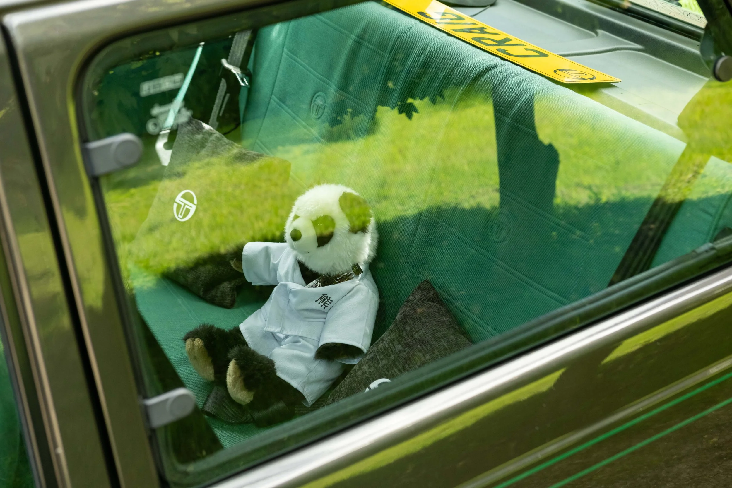 A plush panda bear wearing a white shirt inside a vehicle, seen through the window with grass reflected on the glass, and a yellow caution tape on the roof.