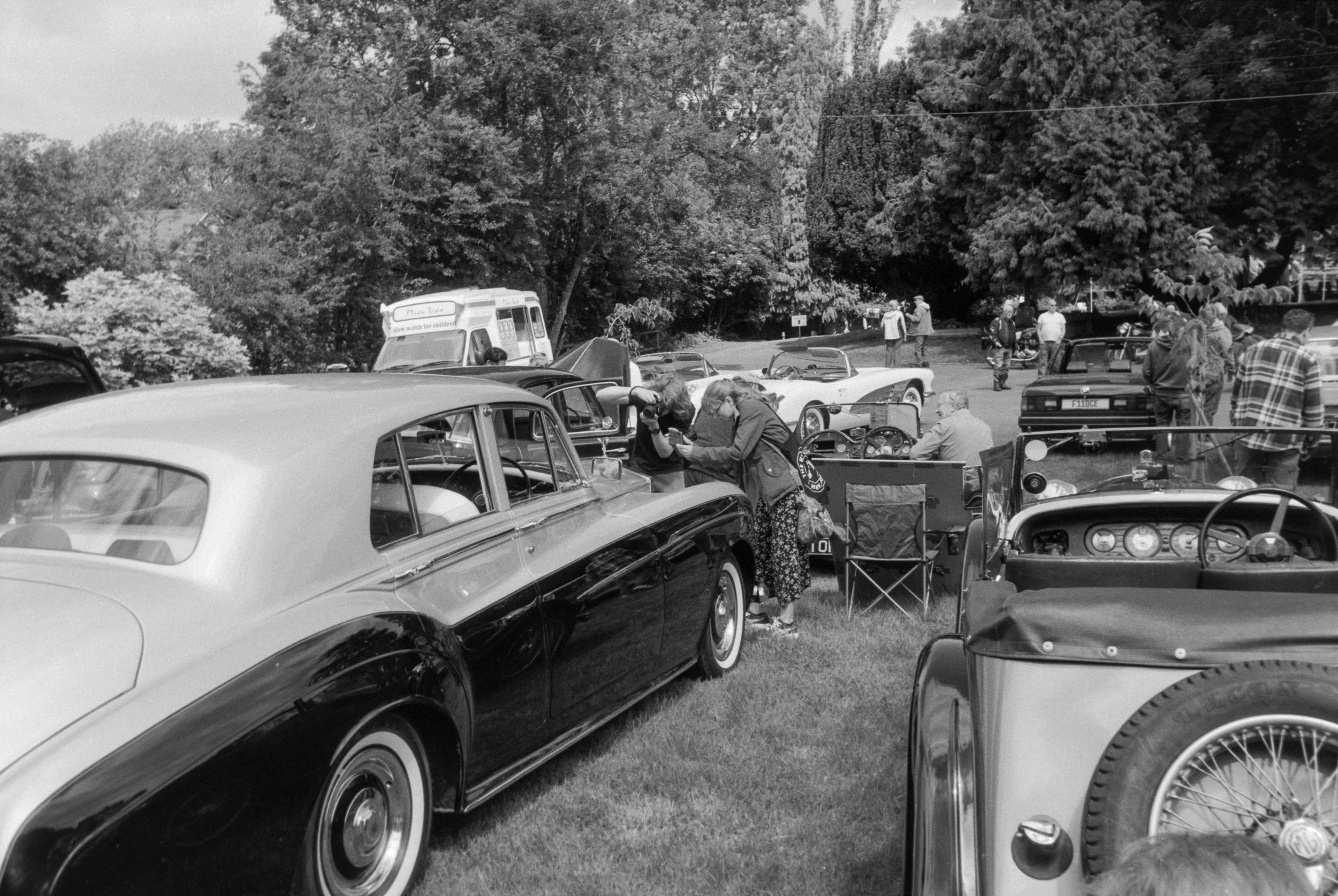A black and white photograph of a gathering of vintage cars parked outdoors, with people walking and socializing among the cars. Trees are in the background, indicating a park-like setting.