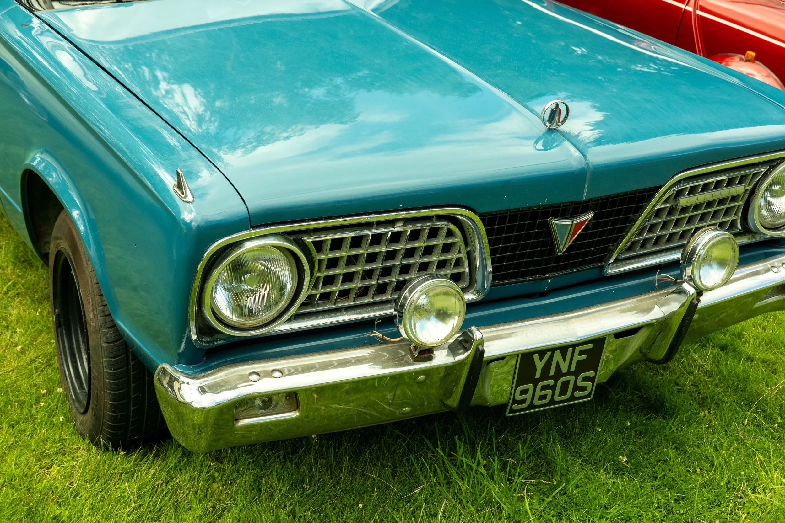 Close-up of a vintage blue car with chrome details, including a grille, bumper, round headlights, and a front license plate that reads 'YNE 960S,' parked on green grass with a red car visible in the background.