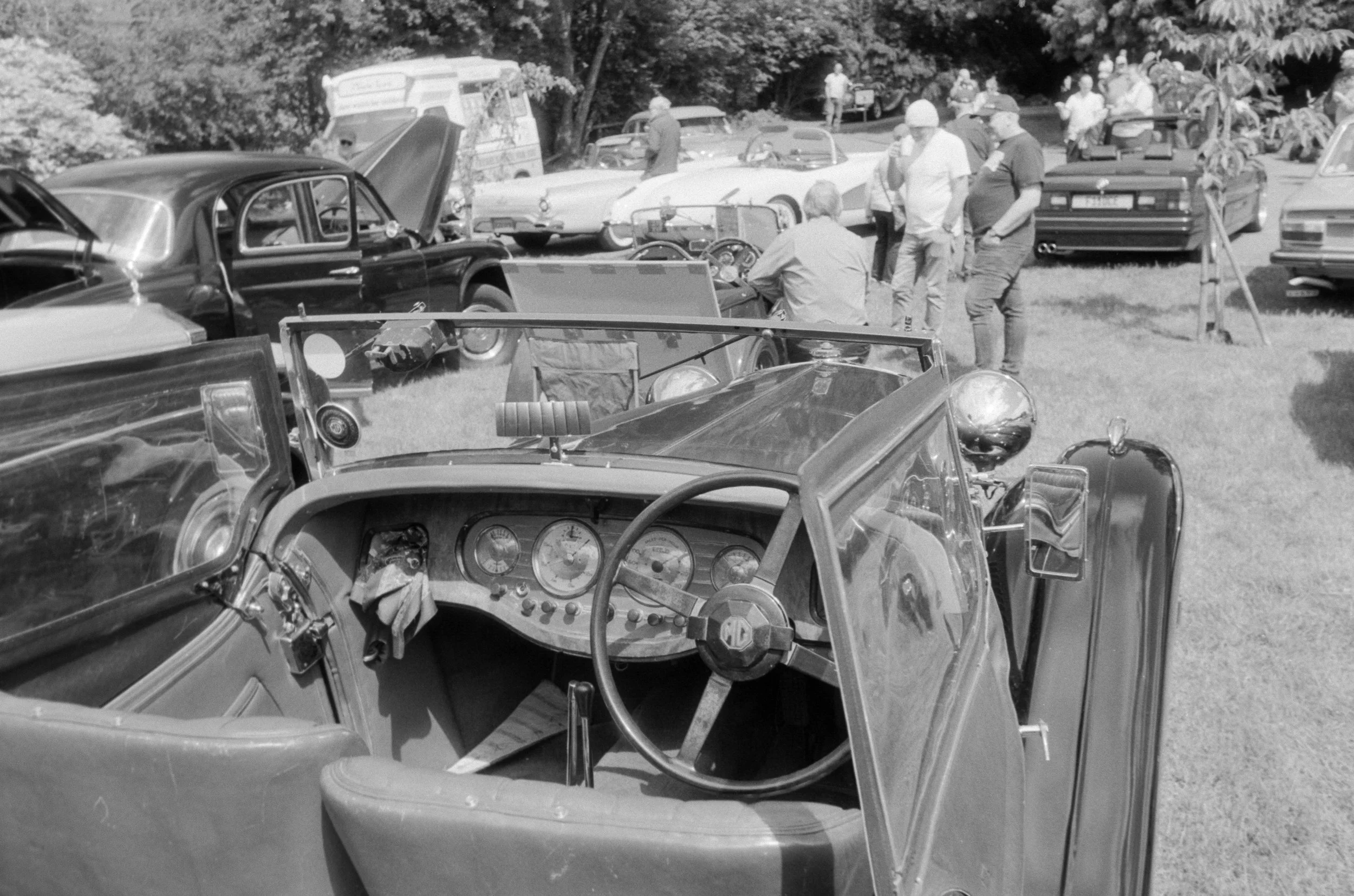 Black and white photo of vintage cars and people gathered outdoors, with some inside or around cars, during what appears to be a car show or gathering.