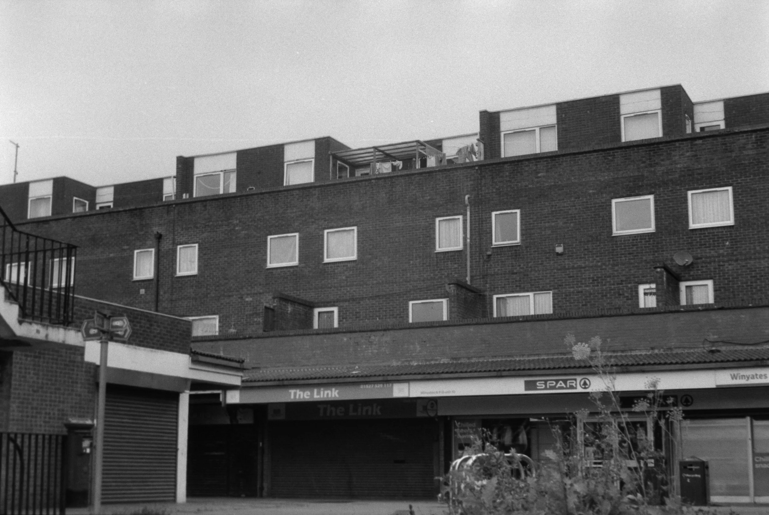 Black and white photo of a multi-story brick apartment building with several windows. On the ground level, there is a storefront with a sign that reads 'SPAR' and 'The Link.' There is a small balcony with laundry hanging outside an upper-floor window