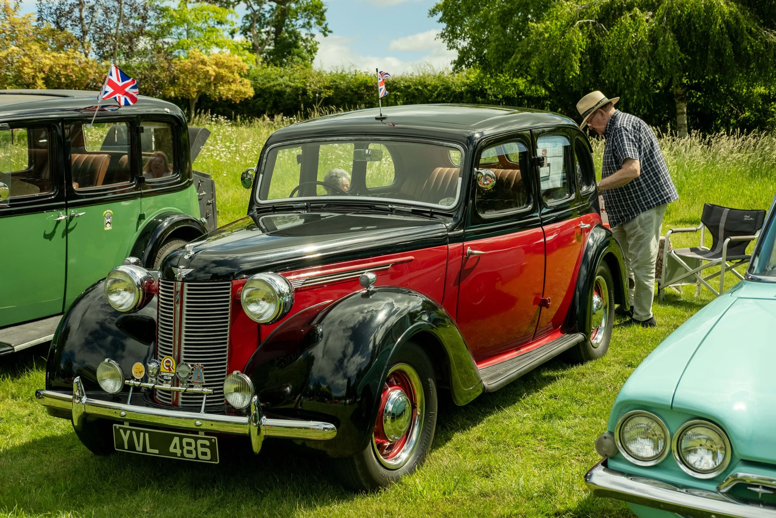 A vintage black and red car parked outdoors on a grassy area with another green vehicle nearby. An elderly man wearing a hat and checkered shirt is inspecting the black and red car. Trees and blue sky with clouds are in the background.