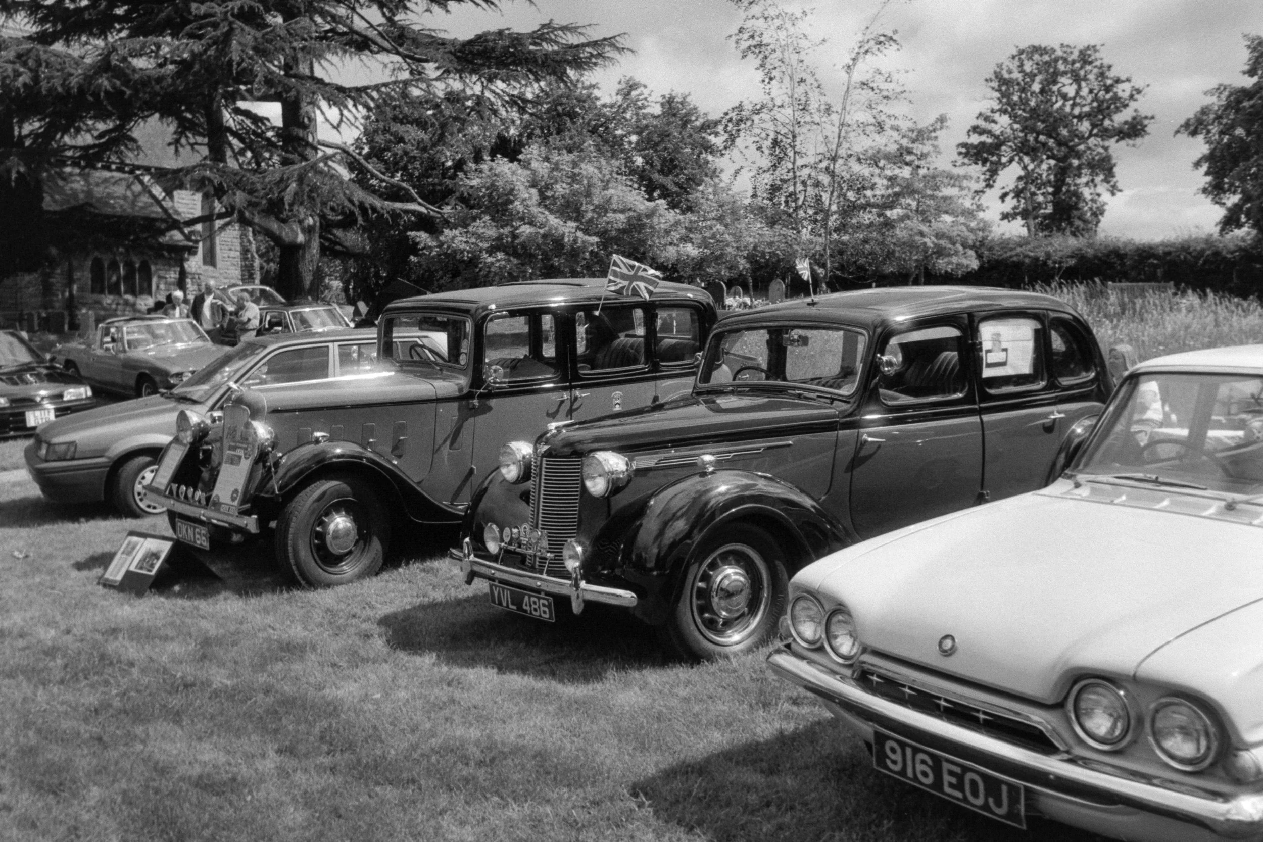 Black and white photograph of a vintage car show outdoors with classic cars on display, some with Union Jack flags, and people gathered around.