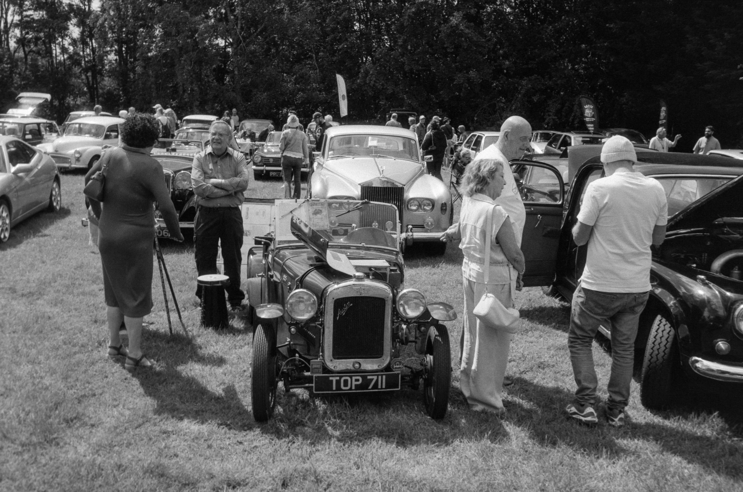 Black and white photo of a vintage car show with people walking around and viewing classic cars in a grassy outdoor area.