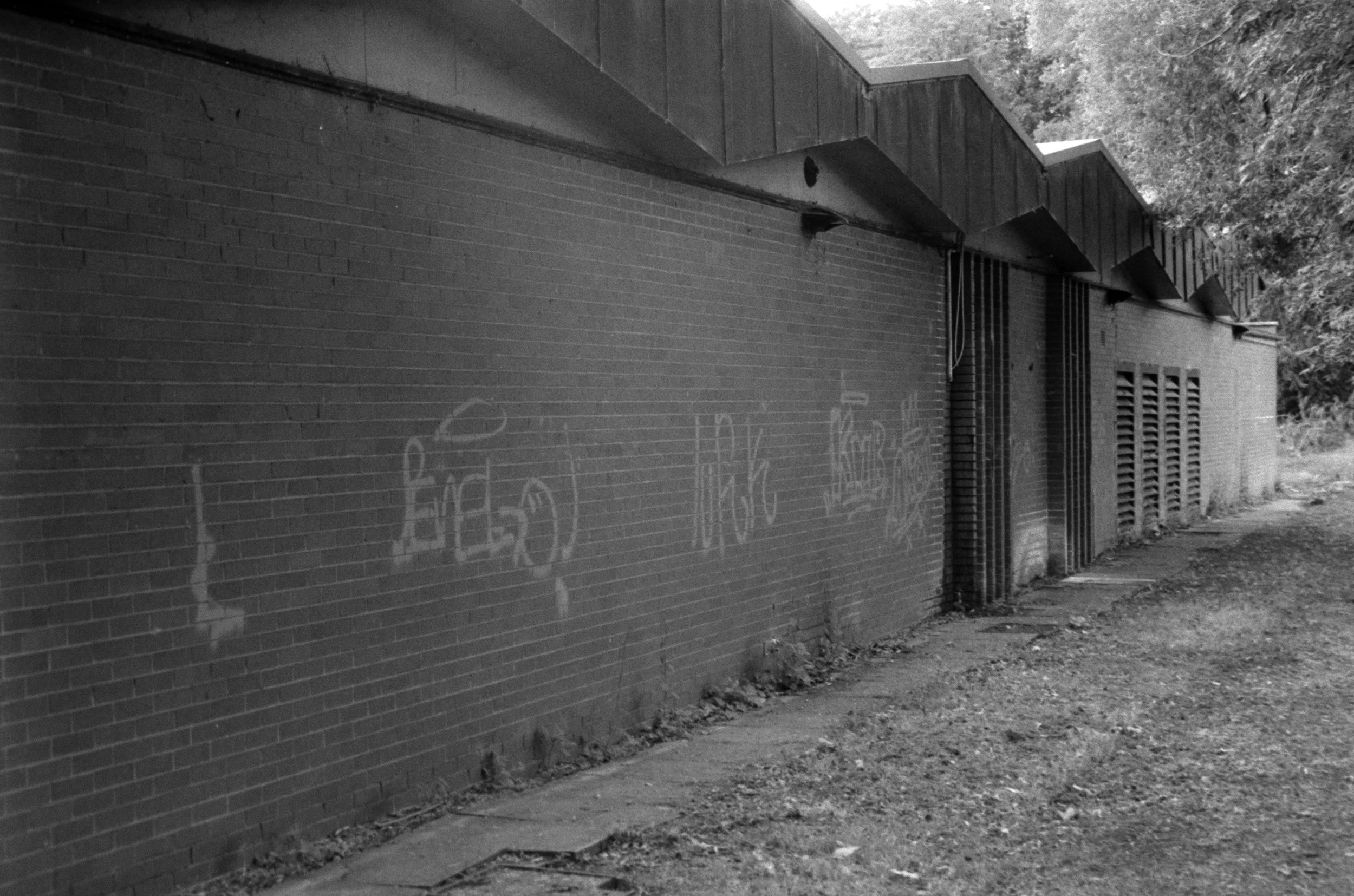 Black and white photo of a brick wall with graffiti and some metal vents, with trees visible above in the background, and a muddy path along the base of the wall.