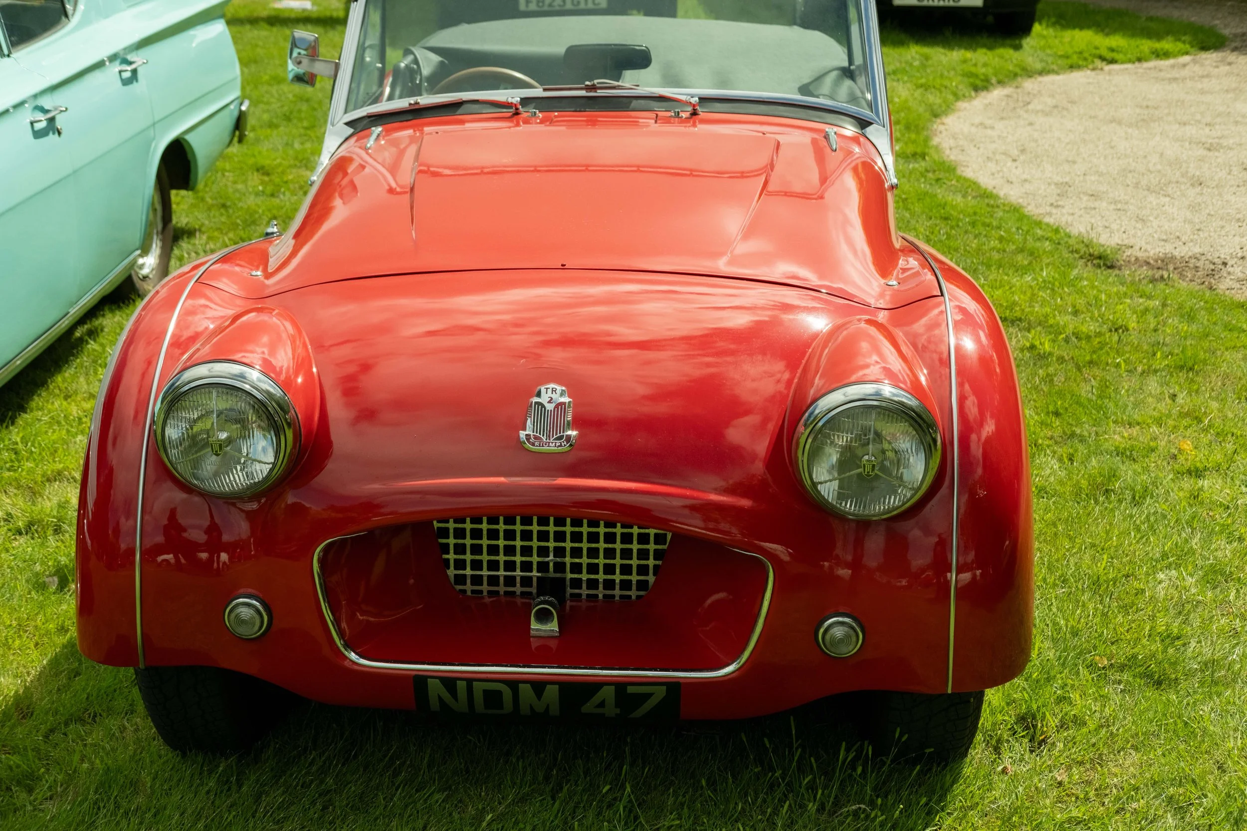 Front view of a vintage red Triumph sports car parked on grass, with another vintage car partially visible on the left.