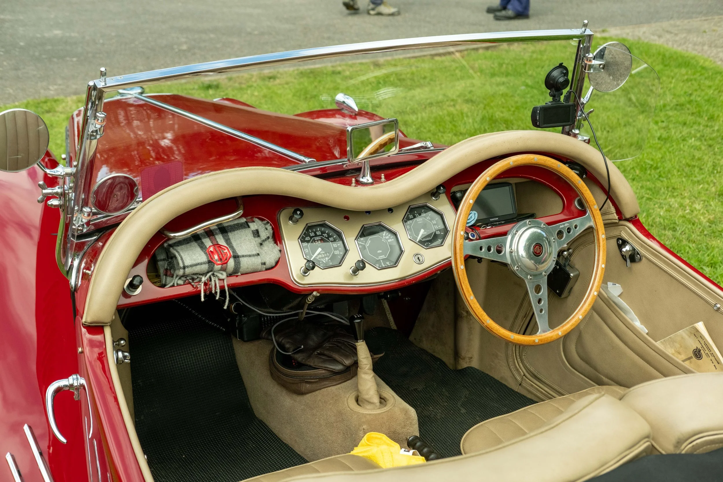 Interior of a vintage red convertible car with a wooden steering wheel, analog gauges, beige leather seats, a windshield with chrome frame, and a grassy background.