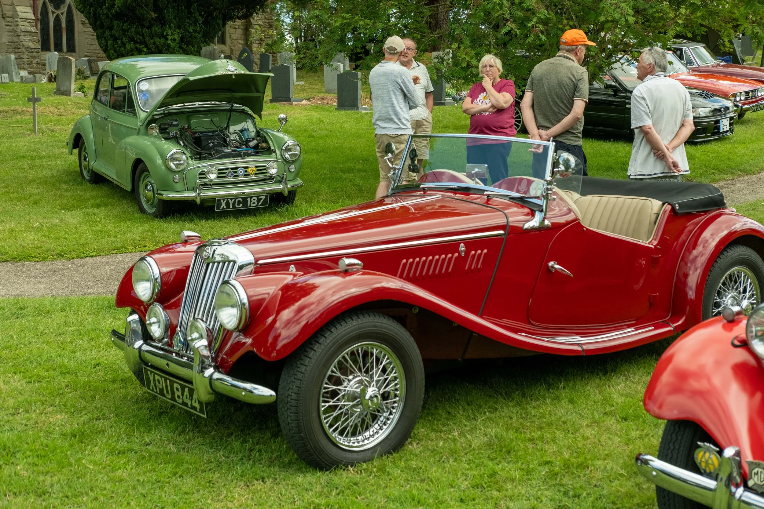 A vintage car show on a grassy field with classic cars including a red MG convertible and a green Mini with its hood open. Several people, mostly middle-aged or older, are walking or talking among the cars, some with arms crossed or hands on hips, un