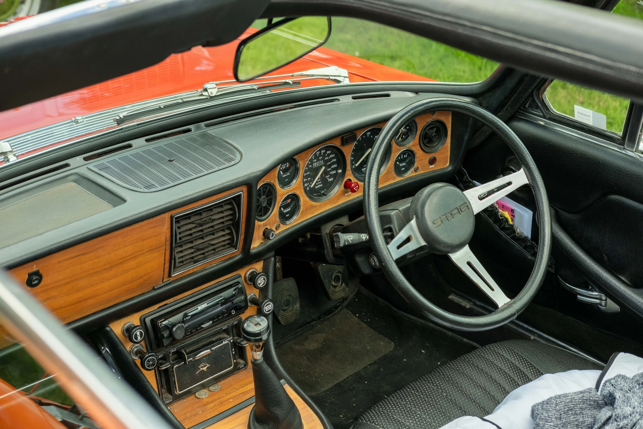 Interior of a vintage car with a wood-paneled dashboard, black steering wheel, and analog gauges, parked on green grass.