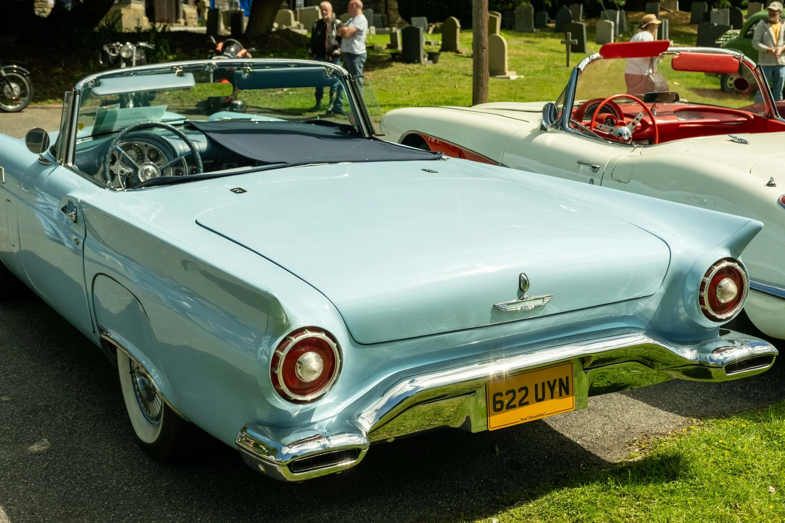 A light blue vintage convertible car parked outdoors, with a black fabric cover over the dashboard, and a red and white vintage car beside it. People are in the background near a grassy area with graves and trees.
