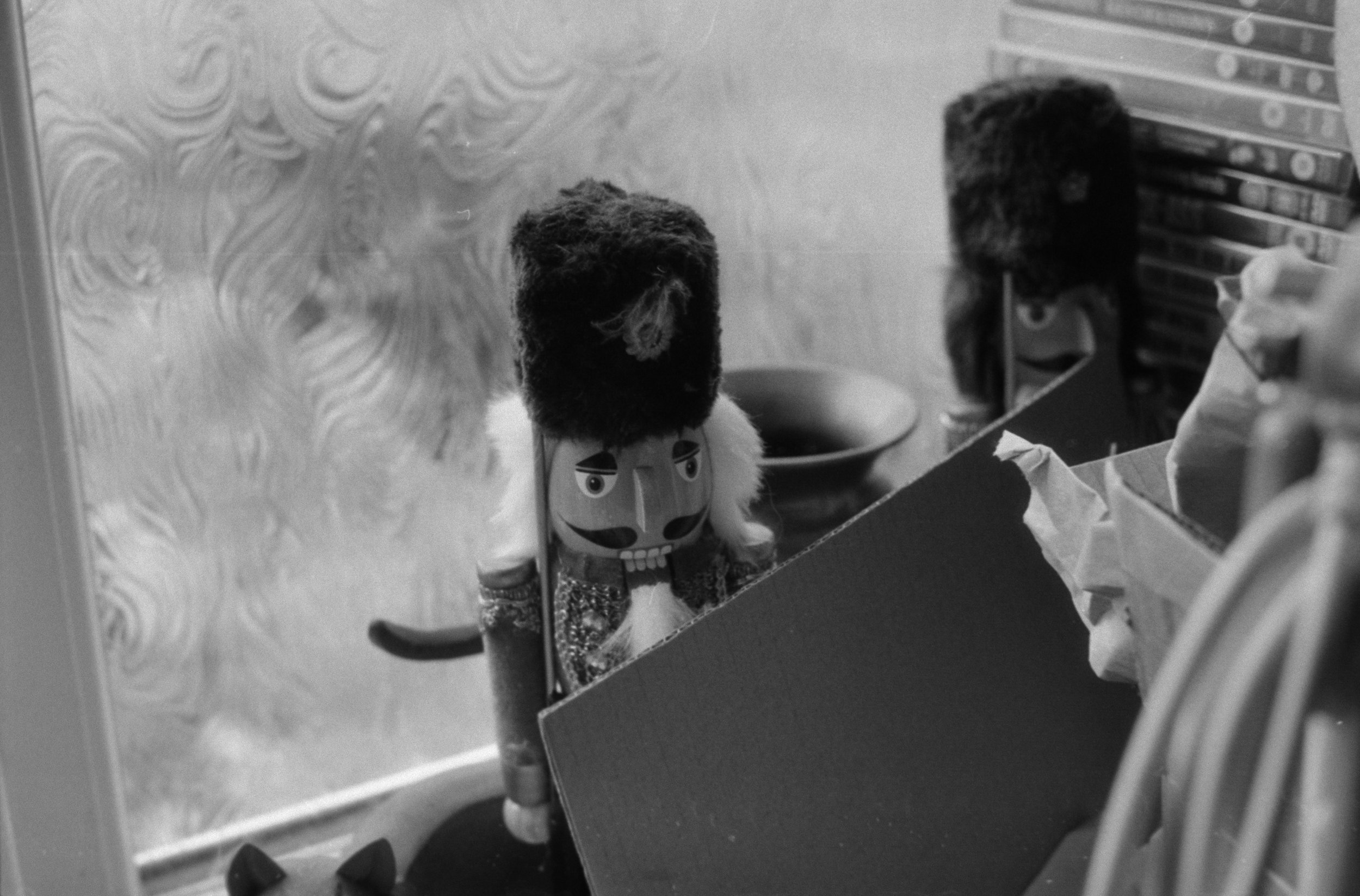 A table with a puppet that has a face with eyebrows and a mustache, wearing a patterned vest, positioned in front of a mirror with an ornate design, reflecting part of the puppet. The background includes shelves with various objects and containers, s