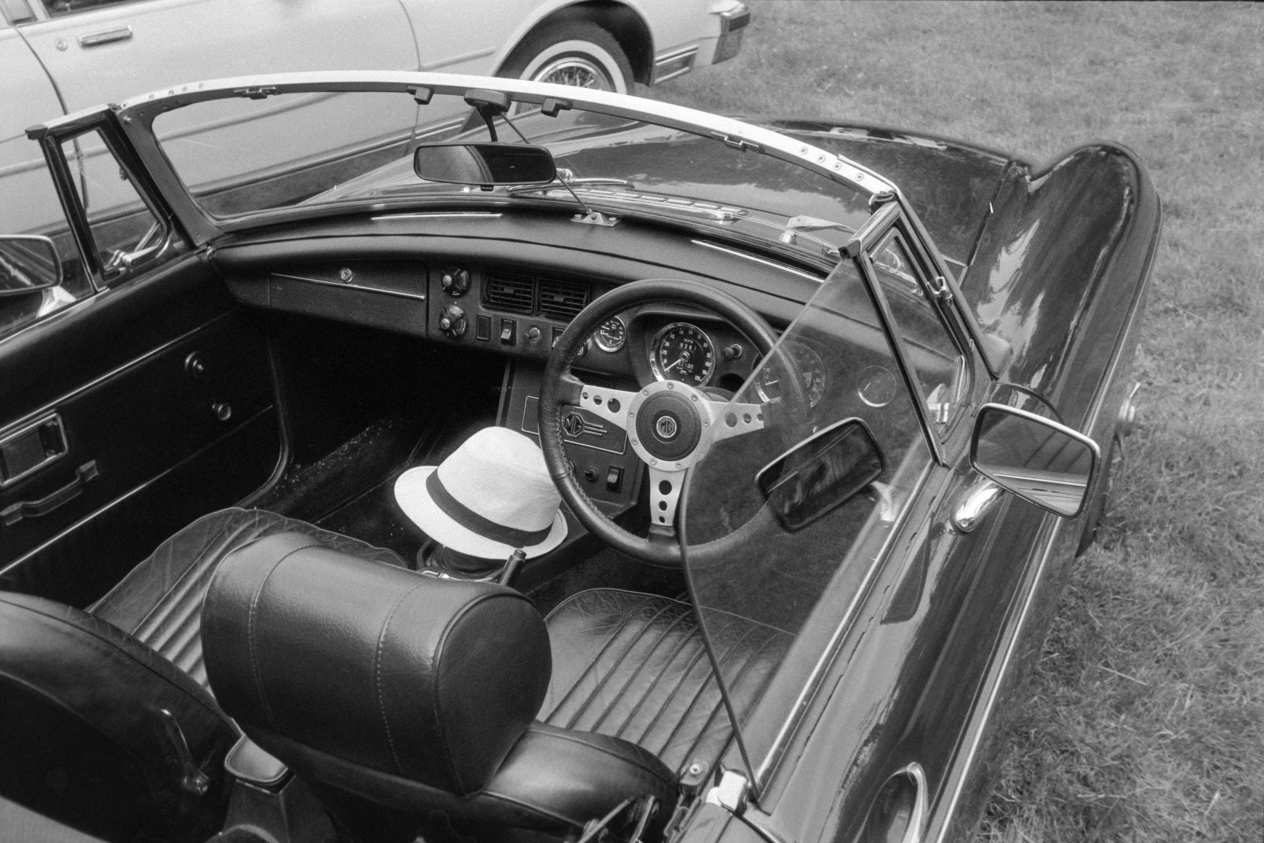 Black and white photo of the interior of a vintage MG convertible car, showing the dashboard, steering wheel, windshield, a straw hat resting on the passenger seat, and a passenger’s leg visible. The car is parked outdoors on grass, with parts of ano