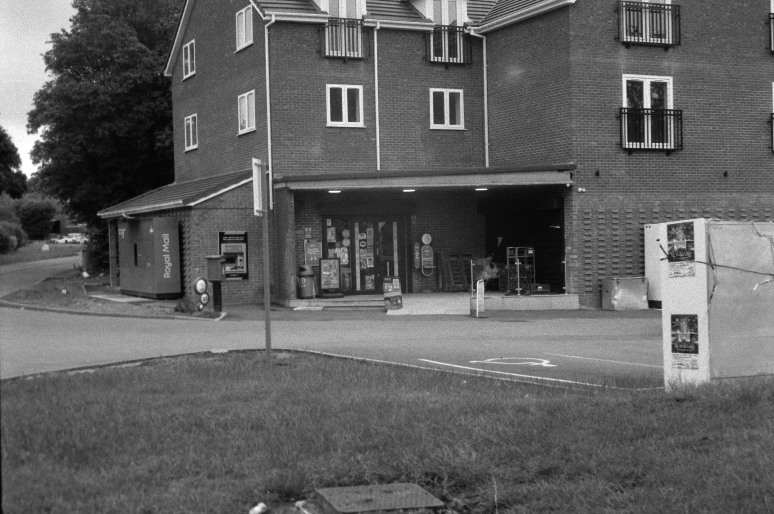 A black and white photo of a brick apartment building with small balconies, and a storefront on the ground floor. The storefront has signage that reads 'Royal Mail' and various posters and signs visible through the window. In front of the building, t