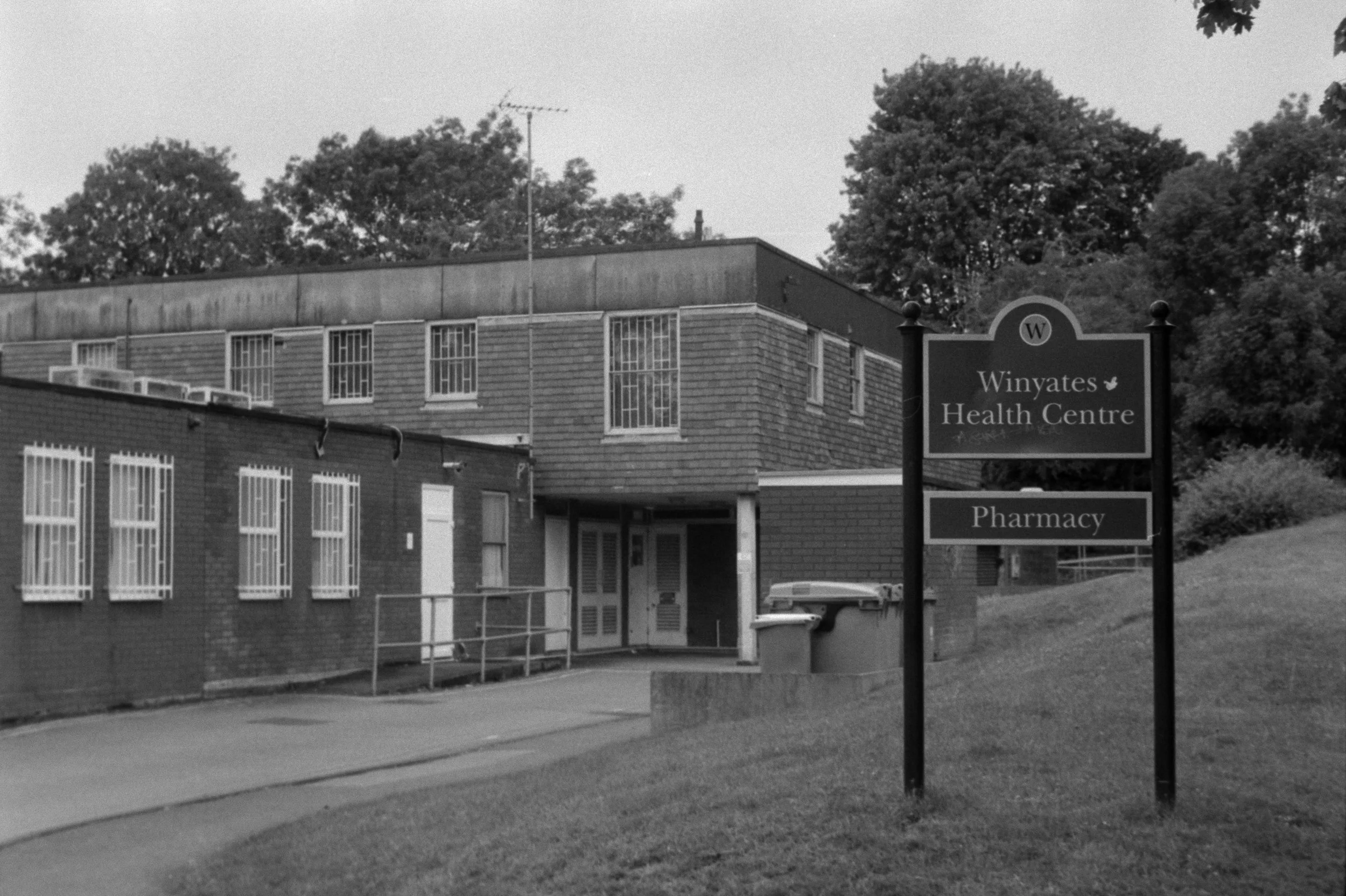 A black and white photo of a building with a sign in front that reads 'Winyates Health Centre' and 'Pharmacy'. There are trees in the background.