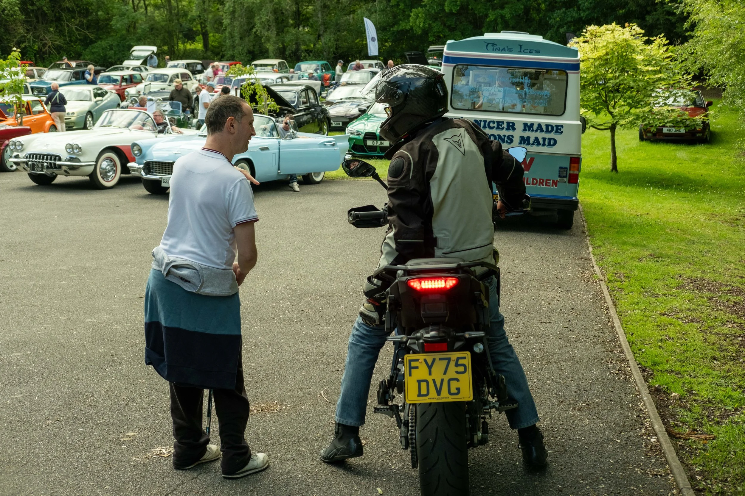 A man and a woman are at a vintage car show, talking next to a motorcycle with a yellow license plate. In the background, several classic cars are parked, and there is a food truck named 'Tina's Ices' and some trees with green foliage.