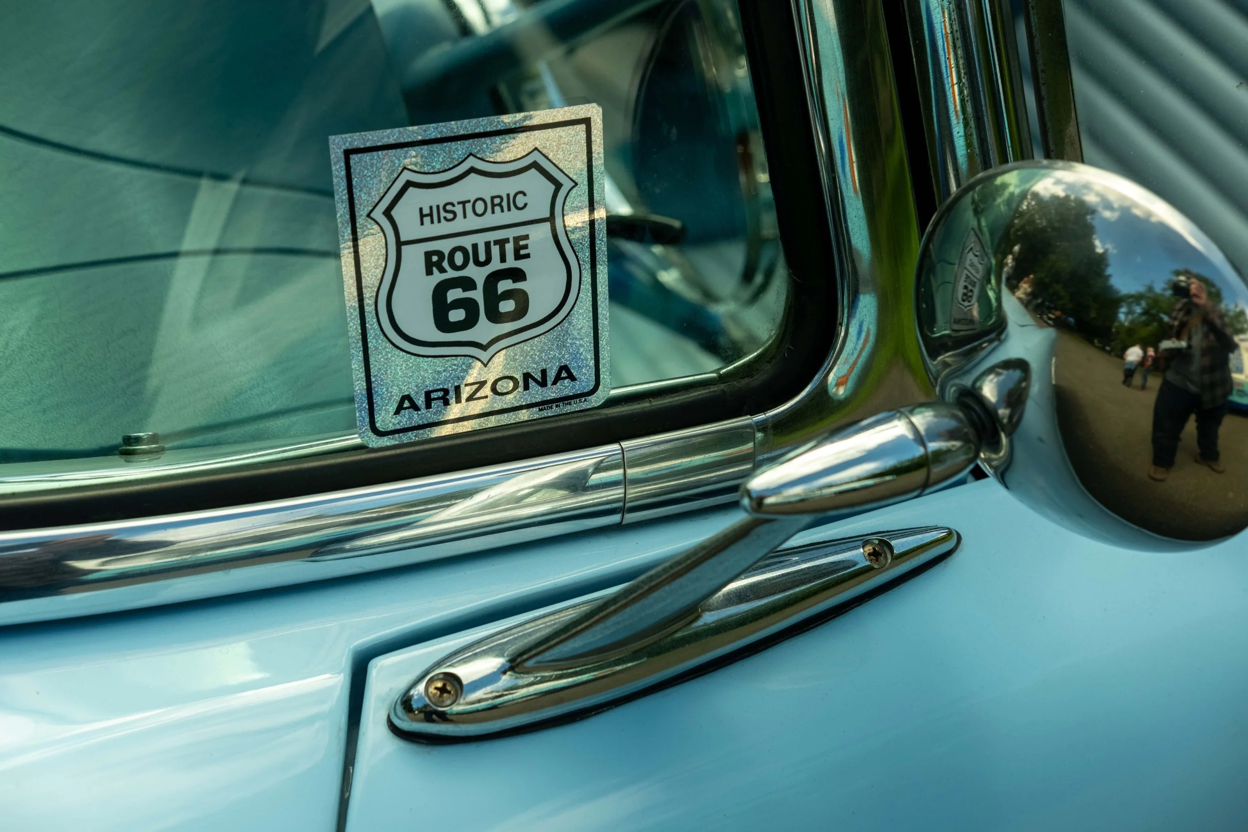Close-up of a vintage car's front with a sticker on the windshield that reads 'Historic Route 66 Arizona,' showing reflections of trees and people in the chrome details.